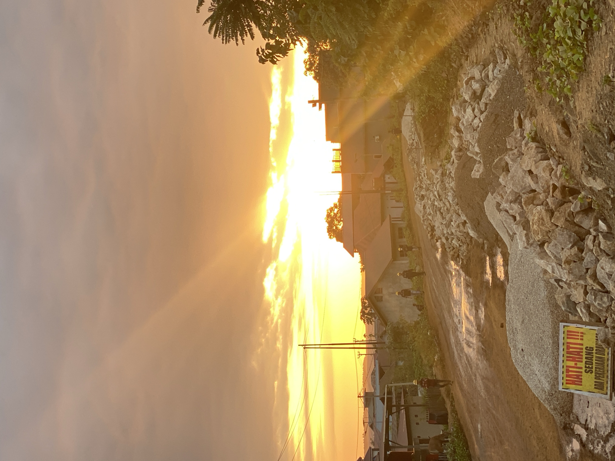 The image depicts a construction site on a dirt road during sunset in an urban or semi-urban area of Indonesia. The sky is filled with warm orange and yellow hues from the setting sun, with prominent sun rays piercing through the clouds. In the foreground, there are piles of gravel and construction materials, along with a yellow warning sign with Indonesian text that reads "HATI-HATI!!!" which translates to "BE CAREFUL!!!". A dirt road, wet in patches, winds upwards, showing signs of recent excavation or road work. Several buildings, appearing to be houses and a commercial structure, are visible in the background. A few individuals, some wearing hard hats, are present on the road, suggesting ongoing construction activity. Green foliage and trees are scattered throughout the scene, with a large tree on the right side of the frame catching the sunset light. The overall impression is that of a community undergoing development amidst a picturesque sunset.