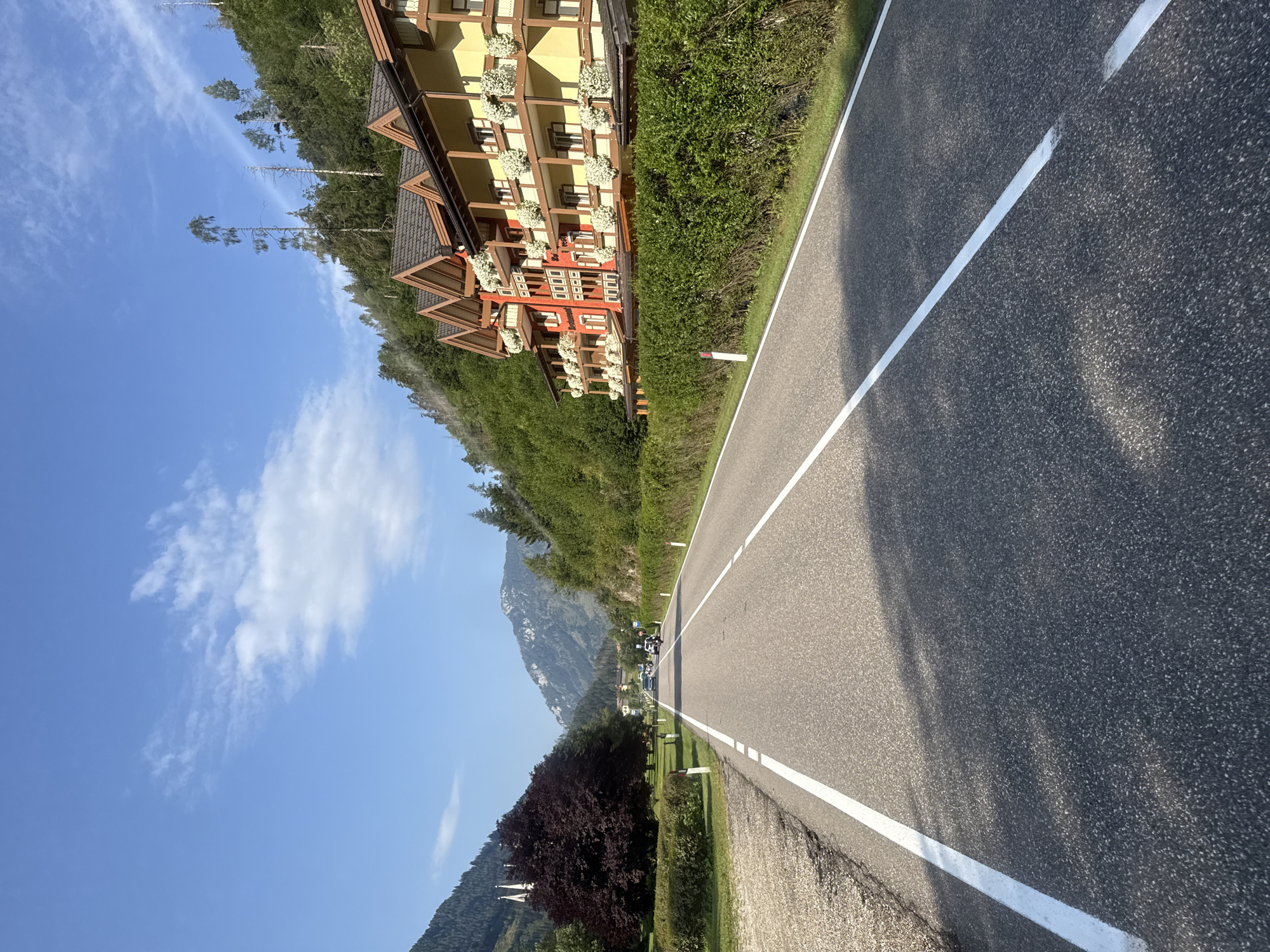 The image shows a paved road with white lane markings, leading into a valley. On the right side of the road is a hotel with red and yellow facades, several balconies with white flowers, and wooden eaves. Behind the hotel, a steep, tree-covered hillside rises sharply. On the left side of the road, there is a grassy area with a dark-leafed tree and what appears to be a small church spire in the distance. The road has a few vehicles on it, including cars and motorcycles, suggesting it is a throughfare. The sky is blue with scattered white clouds. The scene appears to be in a mountainous region, possibly the Dolomites, given the distinctive mountain shapes and architecture. The overall atmosphere is one of a scenic drive through a European mountain town.