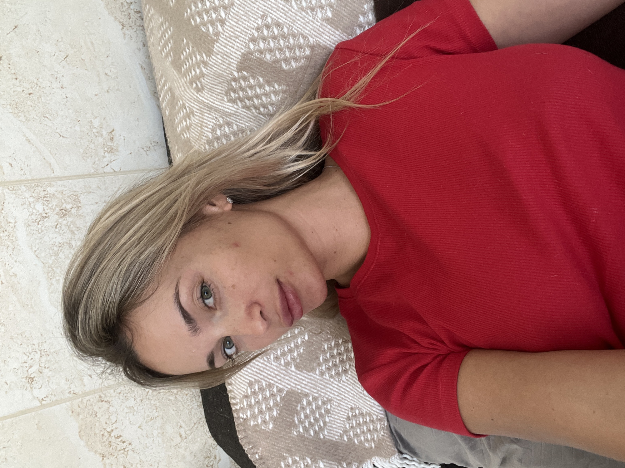 A young woman with blonde hair and green eyes is lying down, her head resting on a patterned pillow. She is wearing a red t-shirt. The background shows a tiled wall. The scene is a close-up shot, focusing on her face and upper body. There is no indication of the city of Fontanka, Ukraine.