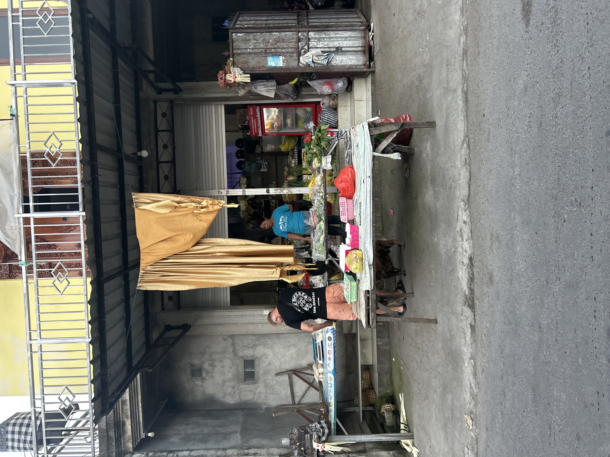A man in a black t-shirt and orange shorts stands at a stall selling produce in Sukawati, Indonesia. He is facing away from the camera, looking down at the items on the table. Beside him, a woman in a blue t-shirt attends to another stall. A small dog can be seen near the man's feet. The stalls are set up outside a shop with a yellow facade and a corrugated metal roof. Various fruits, vegetables, and packaged goods are displayed on the tables. A red Coca-Cola refrigerator is visible inside the shop. The scene appears to be an outdoor market or roadside stand.