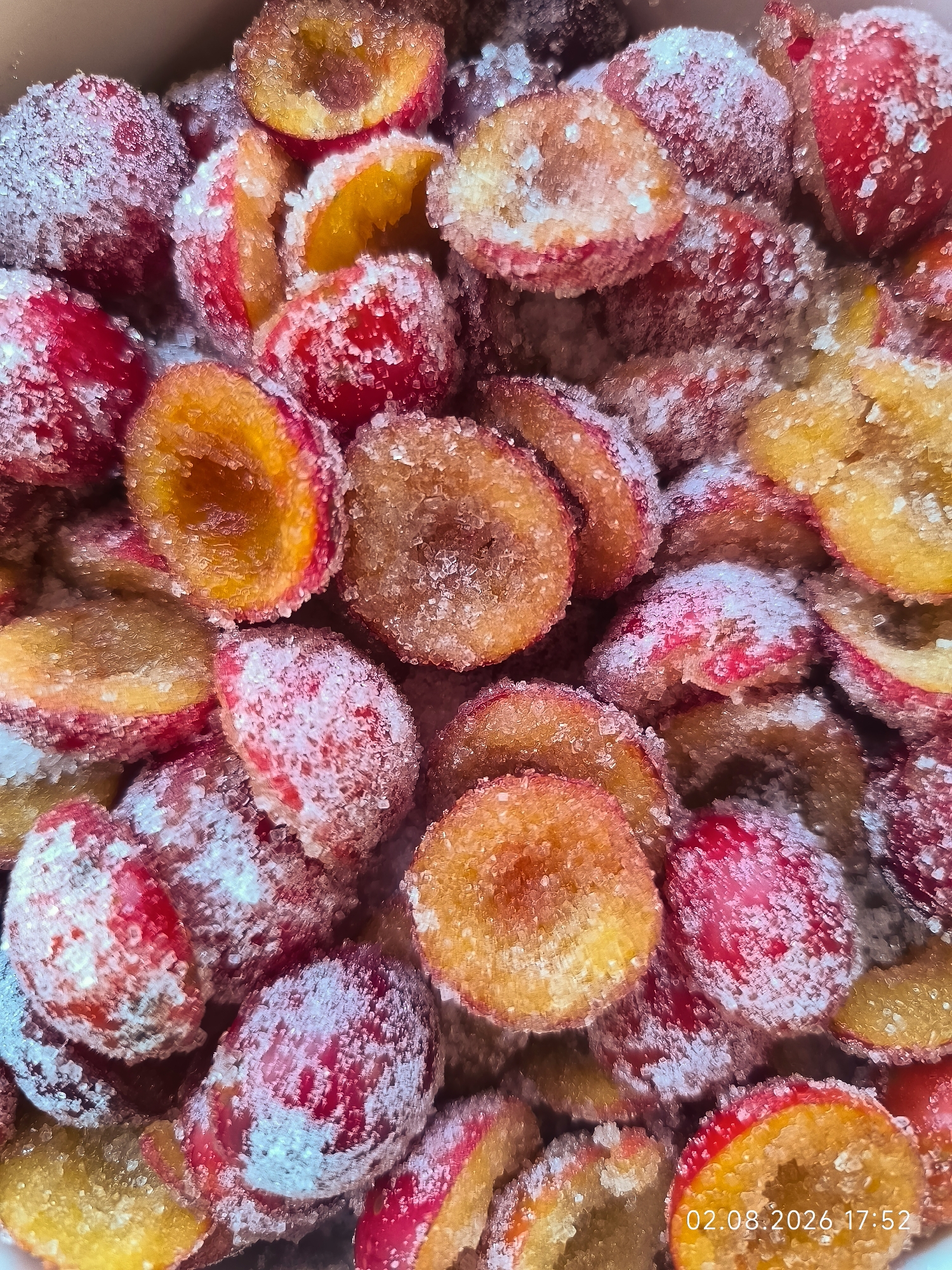The image shows a close-up, overhead view of a pile of cut plums that have been coated in sugar. The plums are halved, revealing their orange-yellow flesh and red skin. The sugar crystals are visible on the surface of the plums, giving them a frosted appearance. The lighting is bright, creating highlights on the sugar and the fruit. The background is out of focus, but appears to be a light-colored container. A timestamp "02.08.2026 17:52" is visible in the bottom right corner of the image. The scene is indoors, likely in a kitchen or food preparation area. There are no people or animals visible in the image.