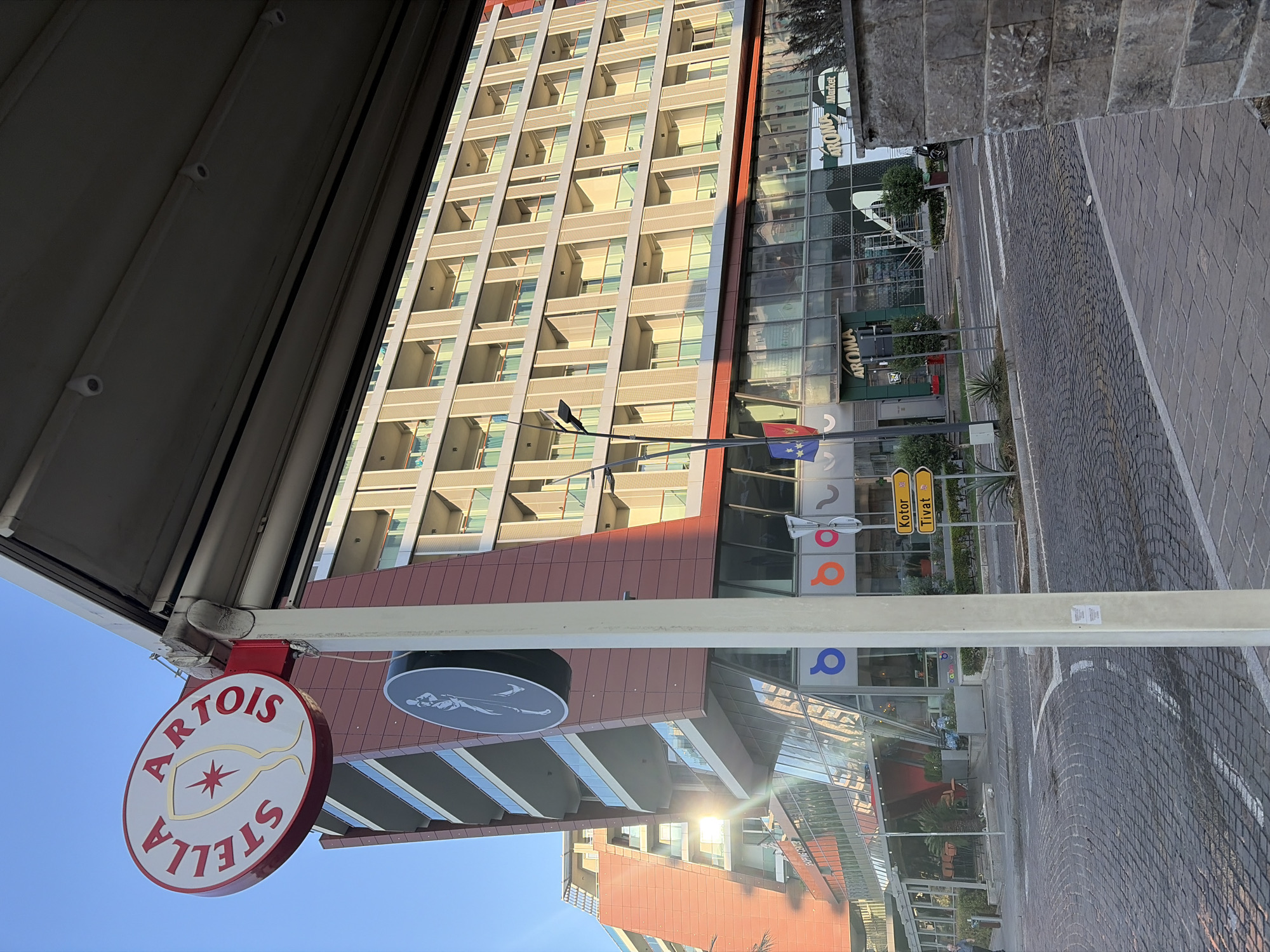 The image depicts a street scene in Budva, Montenegro, on a sunny day. In the foreground, a cobblestone street curves to the left, with a sidewalk made of larger paving stones on the right. A light-colored concrete post stands prominently in the center of the frame. Above the post, a red and white circular sign for Stella Artois is visible. Further down the street, a multi-story building with a modern architectural design, featuring red and cream-colored facades and balconies, dominates the background. The building has large glass windows on the ground floor, revealing shops and commercial establishments, including one labeled "AROMA Market." Two yellow directional signs are mounted on a pole, indicating "Kotor" and "Tivat." The sky is a clear, bright blue. A few trees and shrubs are present along the sidewalk and in front of the building. The overall impression is one of a commercial and urban environment.