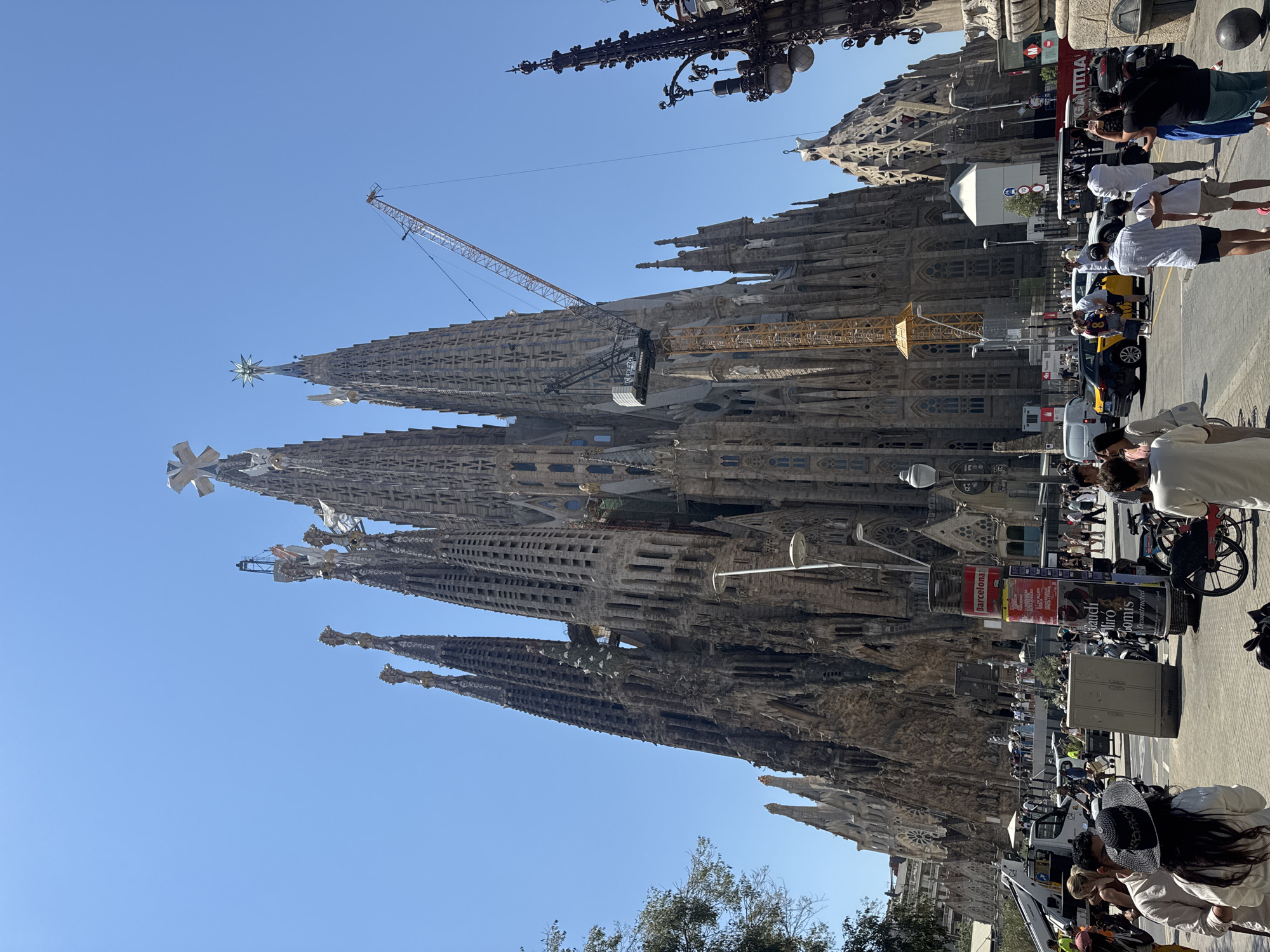 The image shows a wide-angle, low-angle view of the Sagrada Familia in Barcelona, Spain, under a clear blue sky. The iconic basilica is partially obscured by construction cranes, indicating ongoing work. The intricate, Gothic-inspired architecture of the basilica's towers and facades is prominently featured. In the foreground, a bustling street scene unfolds with numerous pedestrians, some walking, others standing and conversing. Various vehicles, including a yellow taxi, a white van, and a red bus, are visible on the road. Several lampposts and a large cylindrical advertisement pillar are also present. The atmosphere appears to be that of a bright, sunny day, with people dressed in summer clothing.