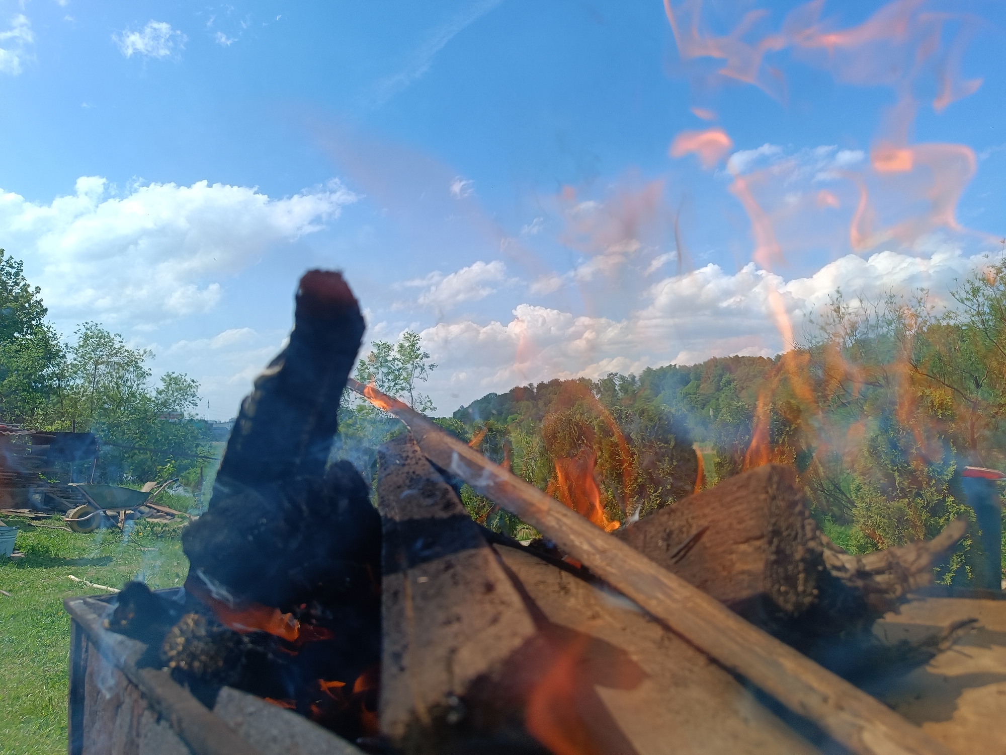 A close-up, low-angle shot captures a fire burning within a metal container. The flames are prominent, flickering and casting orange hues. Charred wood, both large and splintered, lies within the fire, with smoke rising around it.
Behind the fire, a landscape unfolds under a partly cloudy blue sky. Green trees and vegetation cover a gentle slope in the distance. To the left, a section of a green lawn is visible, as well as some structures that appear to be made of wood.
While the image suggests an outdoor setting and rural activity, there is no definitive evidence to pinpoint the location as Motyakovo, Russia. It does however show a woodpile and wheelbarrow near the fire, potentially indicating some kind of yard work is underway. No people are apparent in the frame.