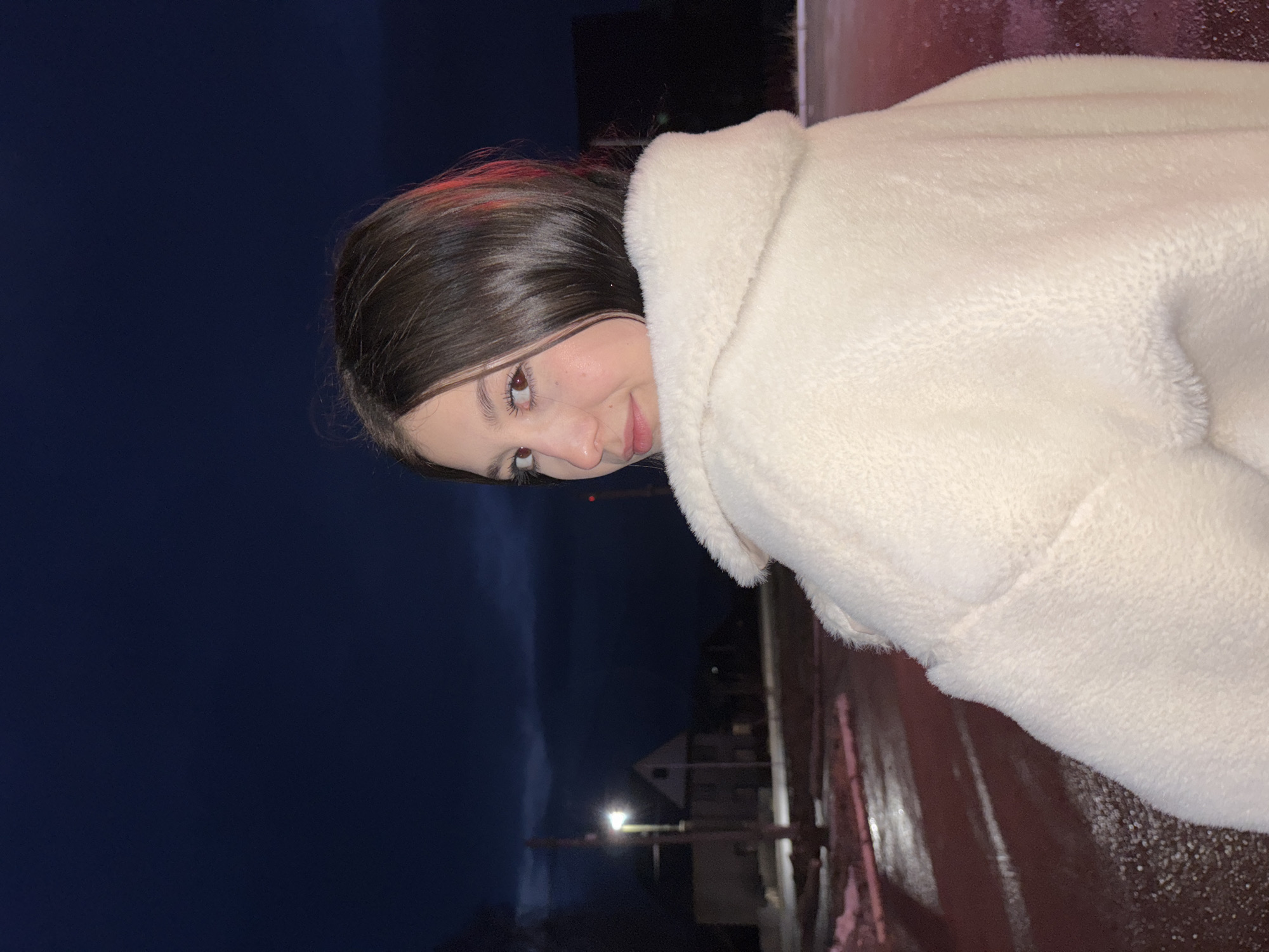A young woman with dark hair and light skin is the main subject of this photo, wearing a fluffy white coat. She is looking at the camera with a slight smile. The background suggests an outdoor setting at night, with a dark blue sky dominating the upper left corner. Streetlights illuminate a building and wet ground, possibly a street. The location appears to be in an urban area, consistent with the city of Фоки, Russia.