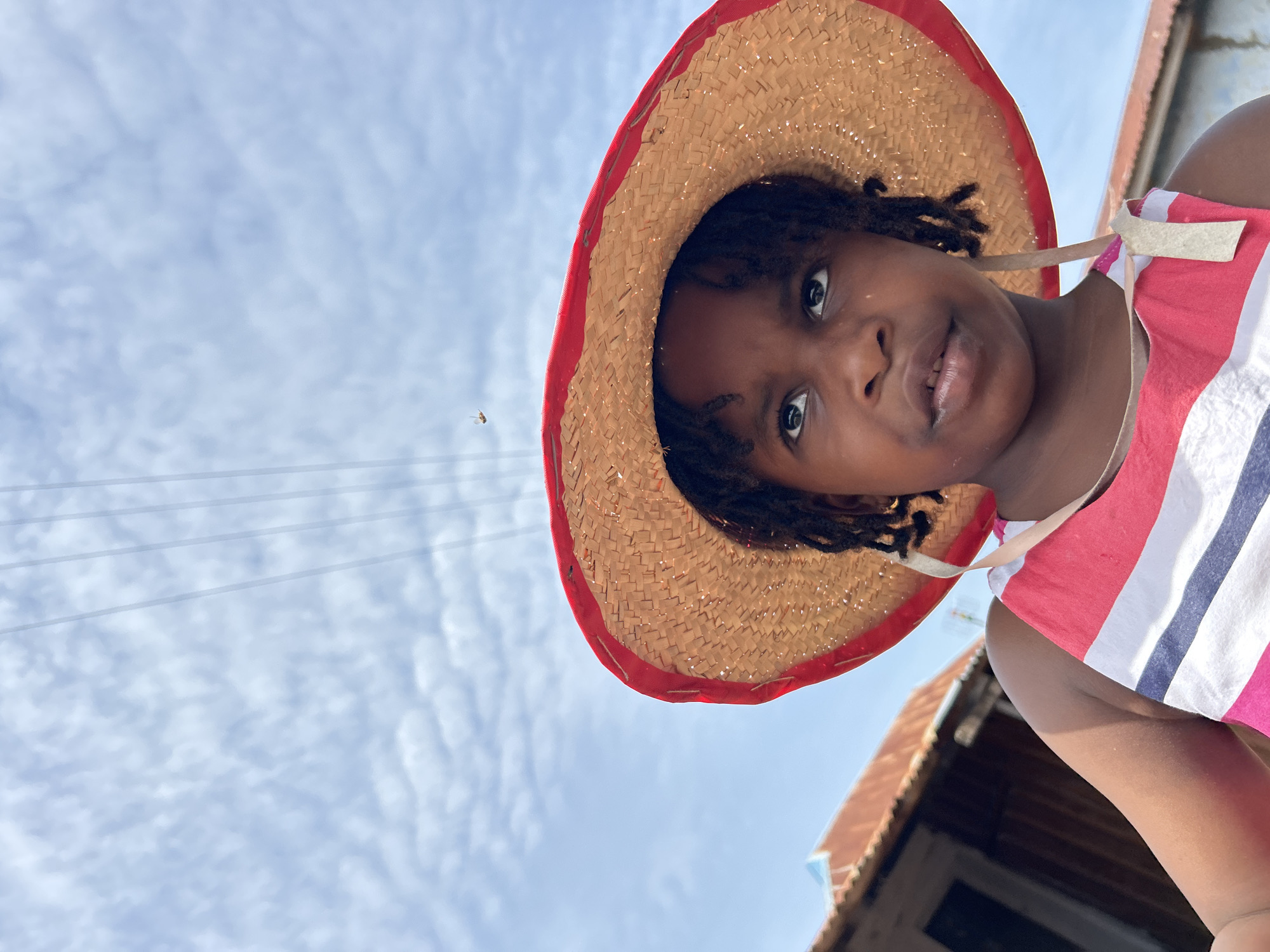 A low-angle, medium shot captures a young Black girl wearing a straw hat with a red brim and a striped tank top. Her eyes gaze upwards, and her mouth is slightly open. The background reveals a cloudy sky, power lines, and the roofs of buildings, suggesting an outdoor setting in Jos, Nigeria. The image is clear and well-lit, focusing on the girl's face and upper body.