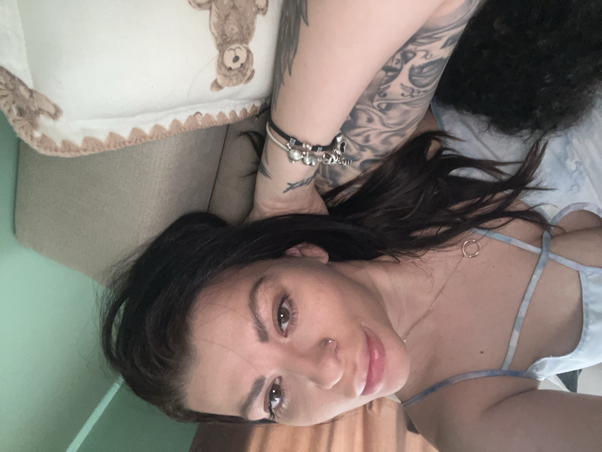 The image shows a close-up, high-angle, selfie-style shot of a woman with dark hair, lying on her side. She is wearing a light blue spaghetti-strap top with a tie-dye pattern. The woman has a nose ring and is wearing a necklace with a circular pendant, as well as a bracelet with charms on her left arm, which also features extensive tattoos. Her left arm is resting on her head. To her right, a pillow with a teddy bear pattern is visible. The background appears to be a sofa or bed. The lighting suggests an indoor setting, possibly during the day. There is no identifiable information indicating the image was taken in Plovdiv, Bulgaria.
