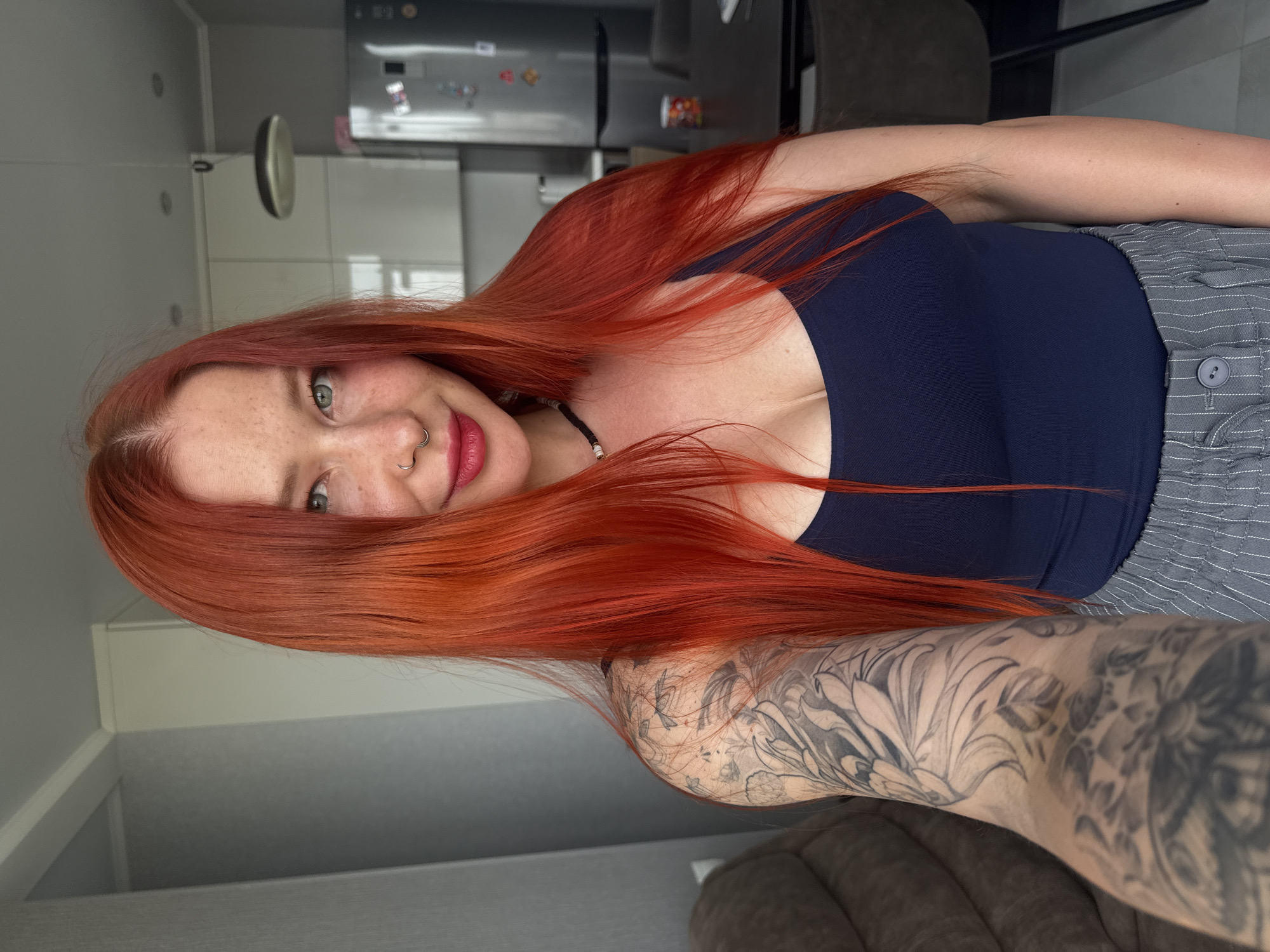 A photograph features a woman with fair skin and bright red hair. She has green eyes and freckles across her face. She is wearing a dark blue, low-cut top. Her left arm, which is heavily tattooed in black ink, is visible. The background shows a modern interior, including a stainless steel refrigerator and a gray kitchen cabinet. The presence of the refrigerator could indicate the location is in Russia as the refrigerators have many magnets.