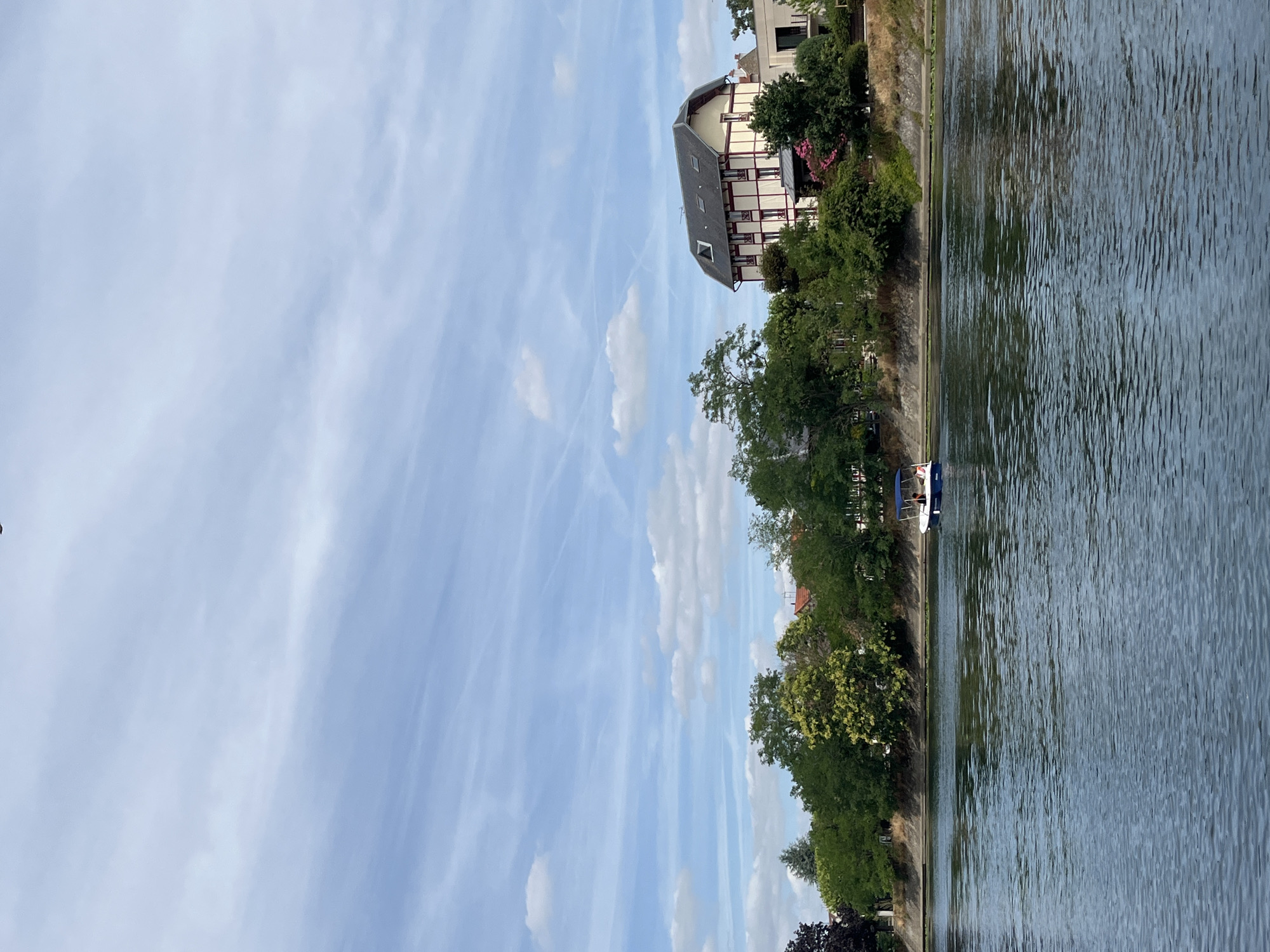 The image depicts a serene riverside scene in Joinville-le-Pont, France. A small boat with two occupants is navigating the calm, blue water of the Marne River. Along the riverbank, lush green trees line the shore, interspersed with a few buildings. Notably, a distinctive half-timbered house with red trim and a dark roof stands out amongst the foliage. The sky above is a pale blue, scattered with wispy white clouds, suggesting pleasant weather. The overall impression is one of tranquility and natural beauty within an urban setting.