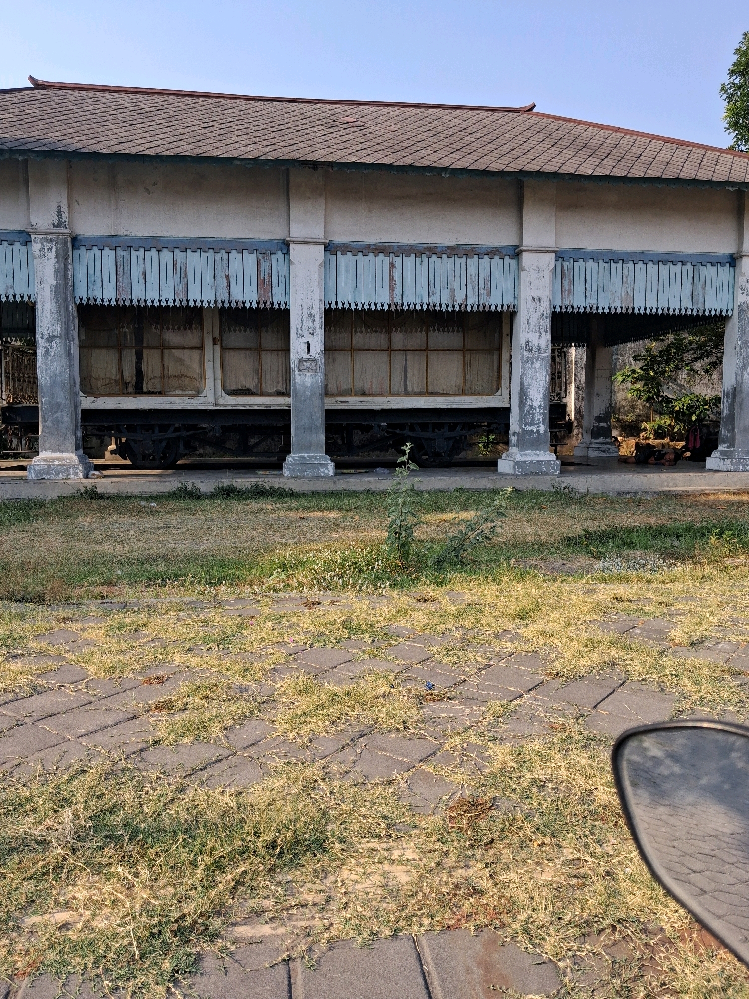 The image captures a scene in Surakarta, Indonesia, likely at the Taman Sari water castle or a similar historical site. In the background, a long, white structure with a dark-tiled roof and white columns stands. Decorative blue wooden panels with a triangular cut pattern are visible along the top facade. Behind these panels, a section of a vintage train car is visible, with its windows covered by light-colored curtains. The train car is situated on tracks in front of the building, which appears to be a sheltered walkway or platform. The ground in front of the building consists of a paved area with square gray tiles, interspersed with patches of dry grass and some weeds. A motorcycle's rearview mirror is visible in the lower right corner of the frame, reflecting the tiled and grassy ground. The sky is clear and blue. The overall impression is of a historical building and train car in a somewhat overgrown, outdoor setting, possibly a museum or tourist attraction.