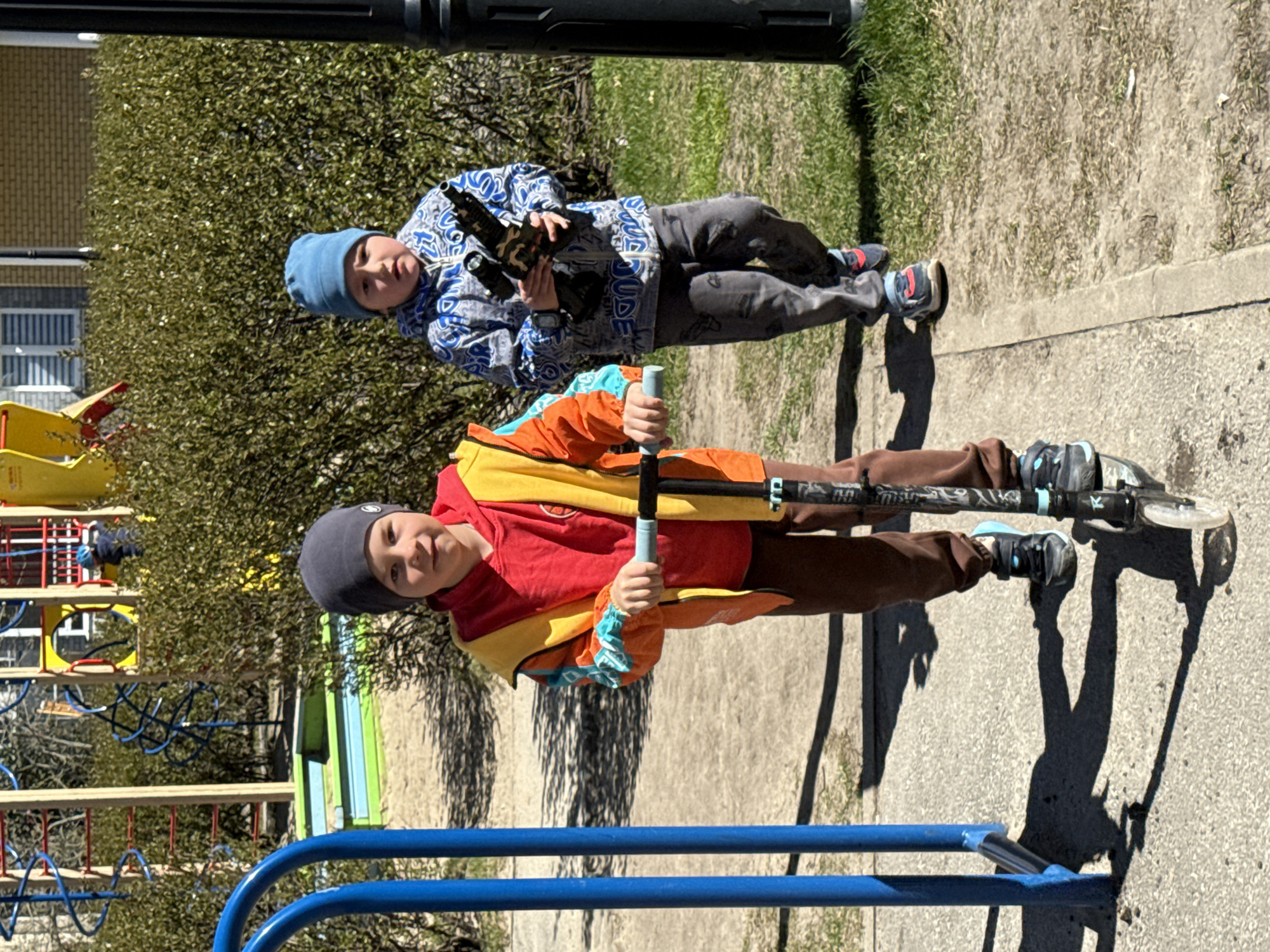 Two young boys are standing outdoors in what appears to be a playground or park. The boy in the foreground is riding a scooter. He is wearing a grey beanie, a red and orange jacket, and brown pants. The second boy, in the background, is holding a toy gun. He is wearing a blue beanie, a patterned jacket, and grey pants. The setting includes playground equipment such as metal bars, and foliage including trees and bushes. A building is visible in the background. The location is Yekaterinburg, Russia.