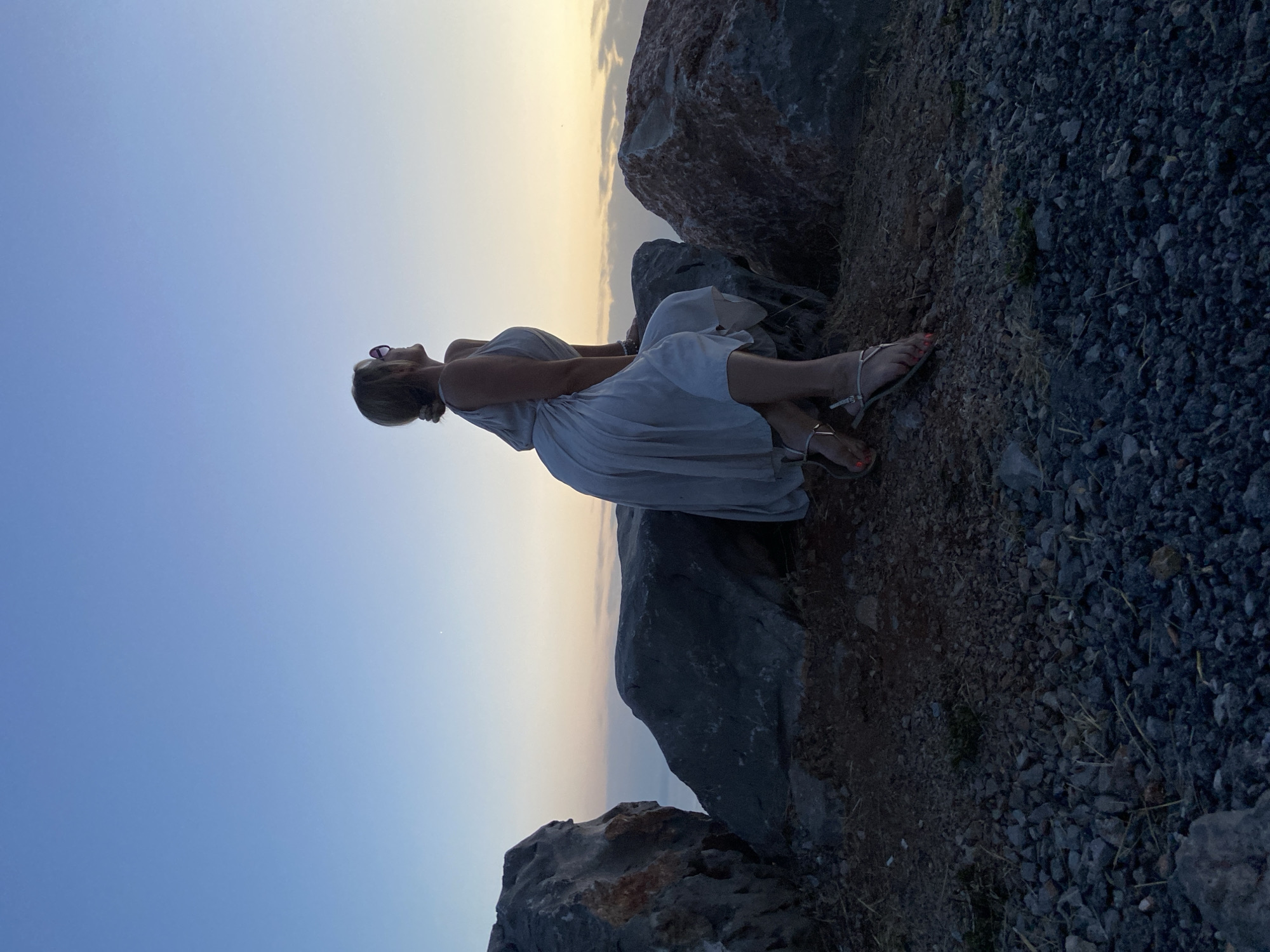 A woman with blonde hair tied up in a bun is sitting on large rocks overlooking a body of water at dusk. She is wearing a light-colored, flowing dress and sandals, and pink sunglasses. The sky is a gradient of blue and orange, indicating sunset. The rocks are dark and rough, with some smaller pebbles and dirt in the foreground. The location appears to be a natural, rocky coastline, possibly near Zagora, Montenegro.