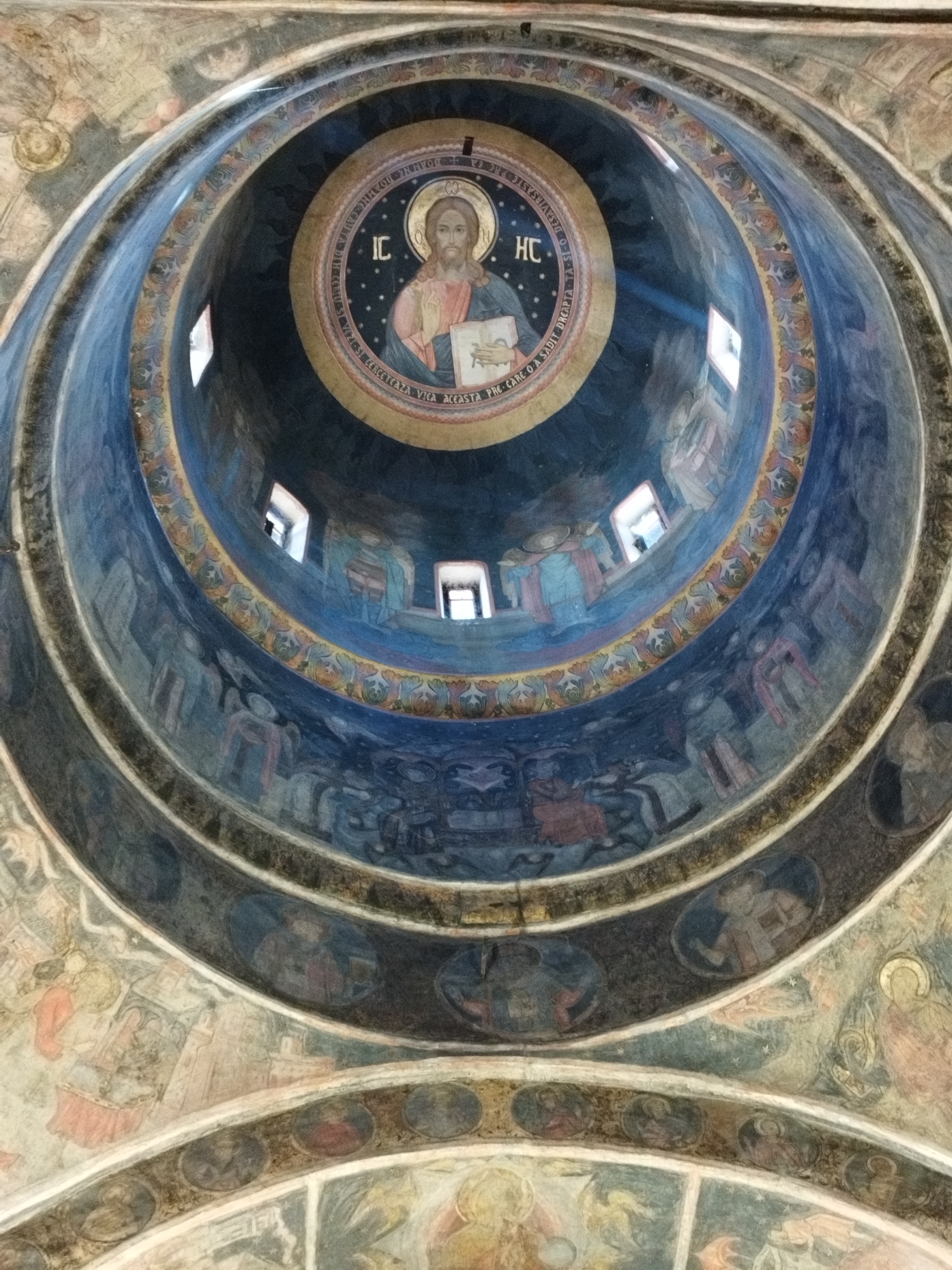 The image shows the interior of a dome within the Stavropoleos Monastery Church in Bucharest, Romania. The dome is adorned with religious frescoes. At the apex of the dome, a circular fresco depicts Jesus Christ holding a book, with a halo and an inscription. Surrounding this central image are concentric rings of decorative patterns and further frescoes of figures, likely saints or biblical characters, rendered in a muted color palette of blues, yellows, reds, and earth tones. Small rectangular windows are visible within the dome's structure, allowing natural light to enter. The overall impression is one of ancient religious art and architecture.