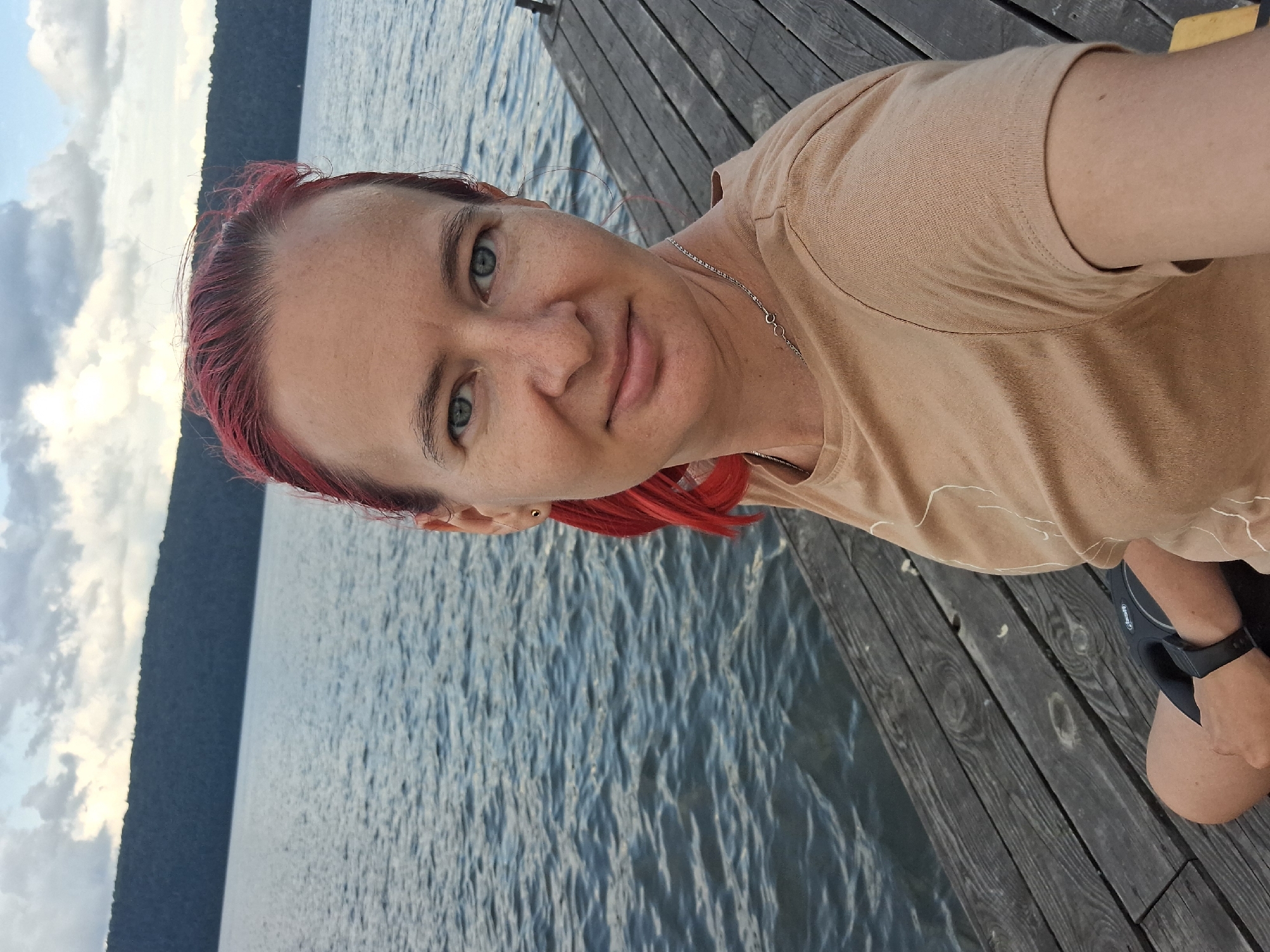 A woman with dyed red hair is sitting on a wooden pier near a body of water. She is wearing a light brown t-shirt and a silver necklace. The water is calm, and a tree-covered hill is visible in the background under a cloudy sky. The pier is made of weathered wooden planks. The scene appears to be a natural outdoor setting, possibly near the town of Атамановка, Russia.