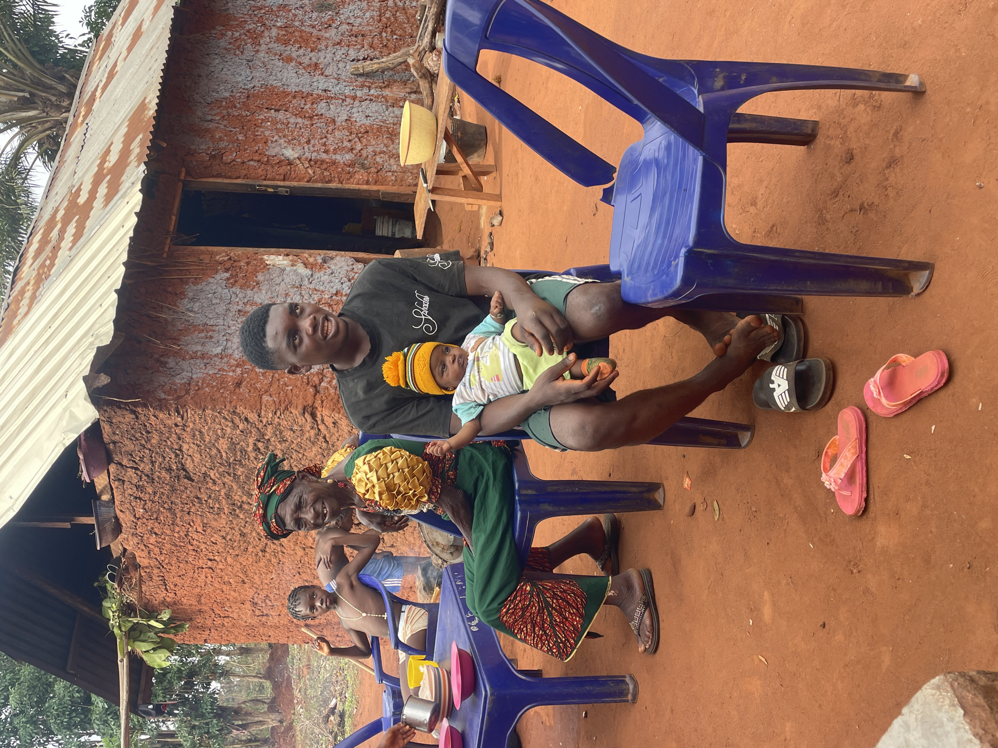 In Ogbe, Nigeria, a group is gathered outdoors in front of a mud-walled structure with a corrugated metal roof. Four individuals are seated on blue plastic chairs around what appears to be a makeshift table. A man in a black shirt sits holding a baby wearing a yellow and orange knit hat. A woman with a headscarf and yellow decorative piece is seated next to him. A young boy sits on a chair beyond her. Various dishes and containers are placed on the blue table. In the background, a clay-colored house with a dark doorway is visible. The ground is reddish-brown. Several pairs of flip-flops rest on the ground.