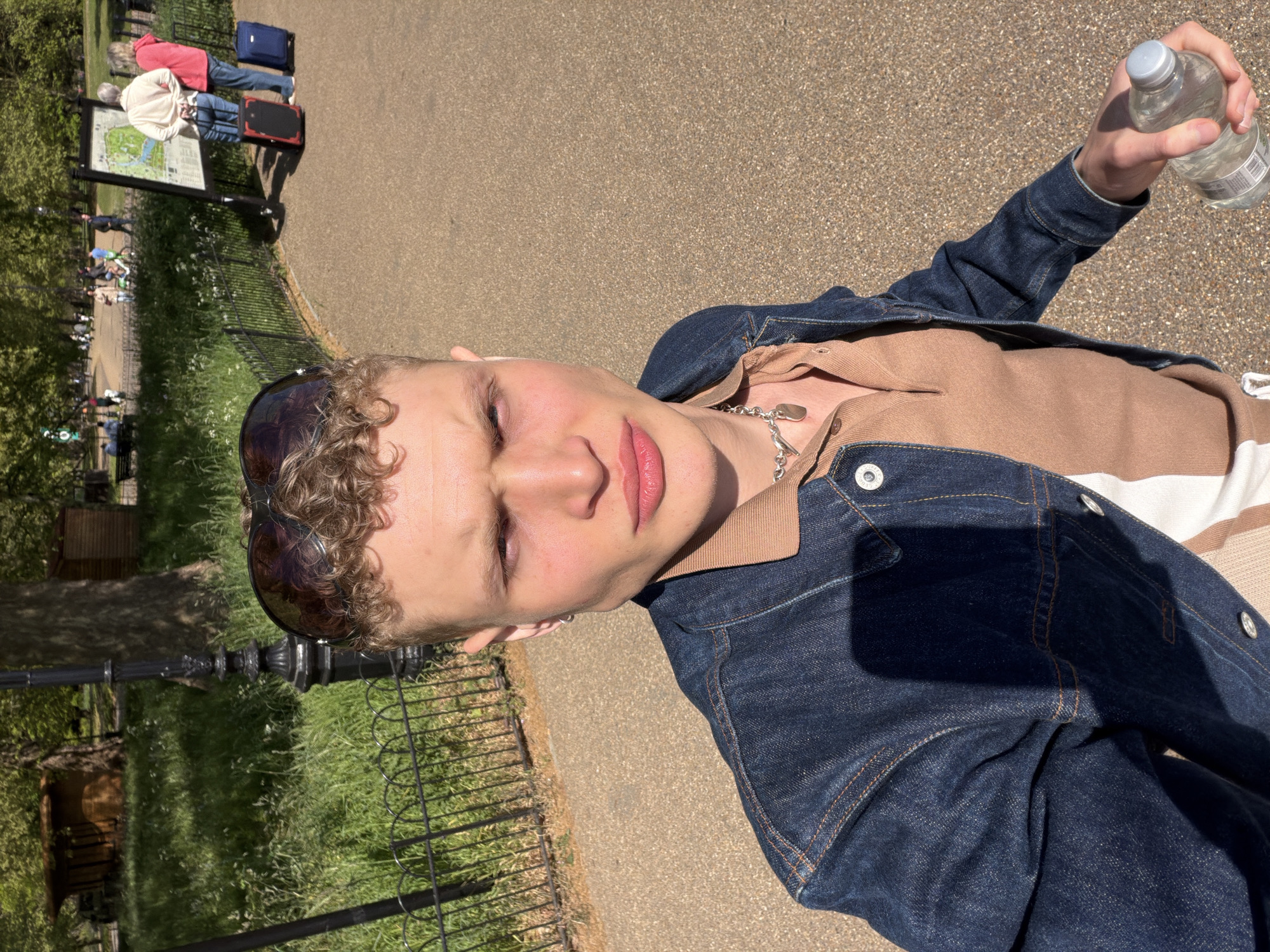 The image shows a person standing on a path, holding a bottle of water. They are wearing sunglasses, a brown shirt with a beige and white striped collar, and a dark blue denim jacket. A silver necklace is also visible.
In the background, there is a grassy area with a low metal fence. People are walking on a path behind the fence, and some are looking at a sign with a map on it. There are trees and other greenery visible.
Based on the sign with the map and the overall look of the surroundings, the location is likely in Hyde Park, London, United Kingdom.