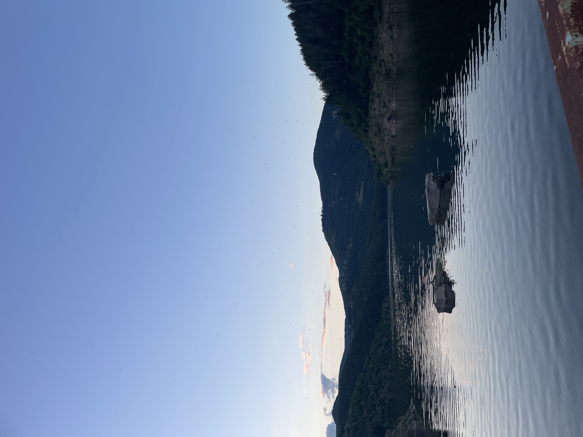 The image depicts a serene lake surrounded by forested hills under a pale blue sky. Several small, dark objects, possibly birds or insects, are scattered across the sky. The water is calm, reflecting the muted colors of the landscape. Two rectangular, light-colored objects protrude from the water in the foreground. The scene appears to be a natural landscape, potentially a reservoir or a large lake, given the presence of man-made structures like the rocky embankment in the bottom right corner which shows signs of wear and rust. The lighting suggests it is either early morning or late evening. The location is identified as being in Suhovo, Bulgaria.