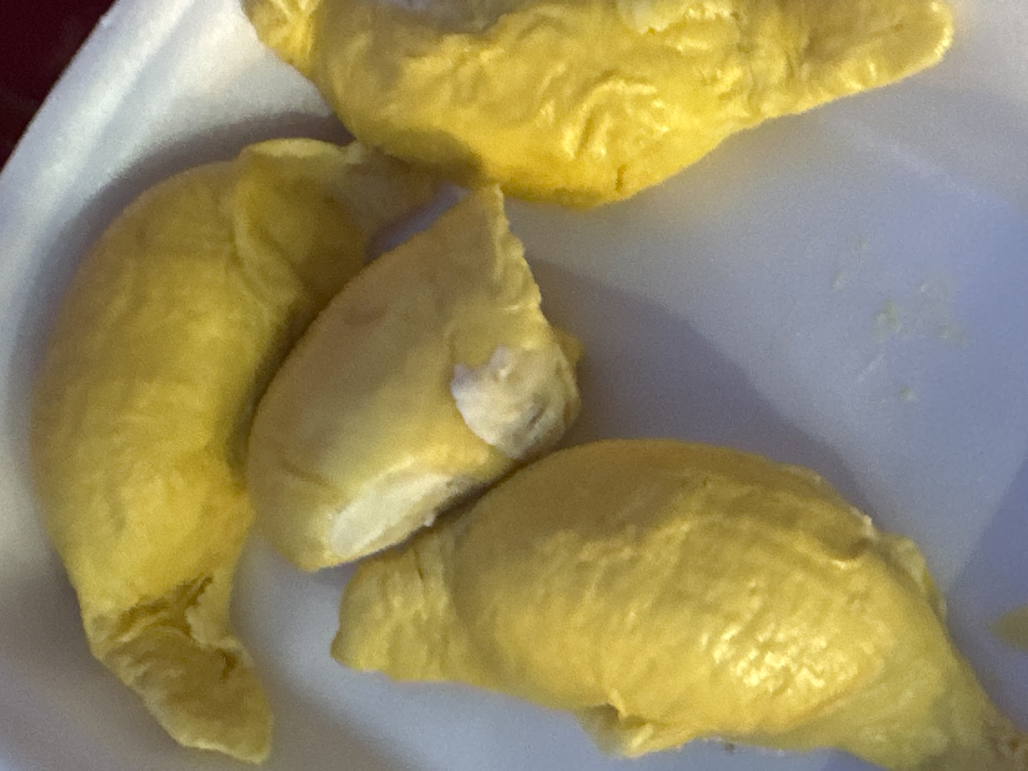 A close-up, overhead shot shows a white styrofoam container filled with several pieces of yellow durian flesh. The durian pieces are irregularly shaped and vary in size. One piece in the upper center of the image has a noticeable white fleshy interior. The background is a blurred, pale purple, suggesting an outdoor setting in an unknown location.