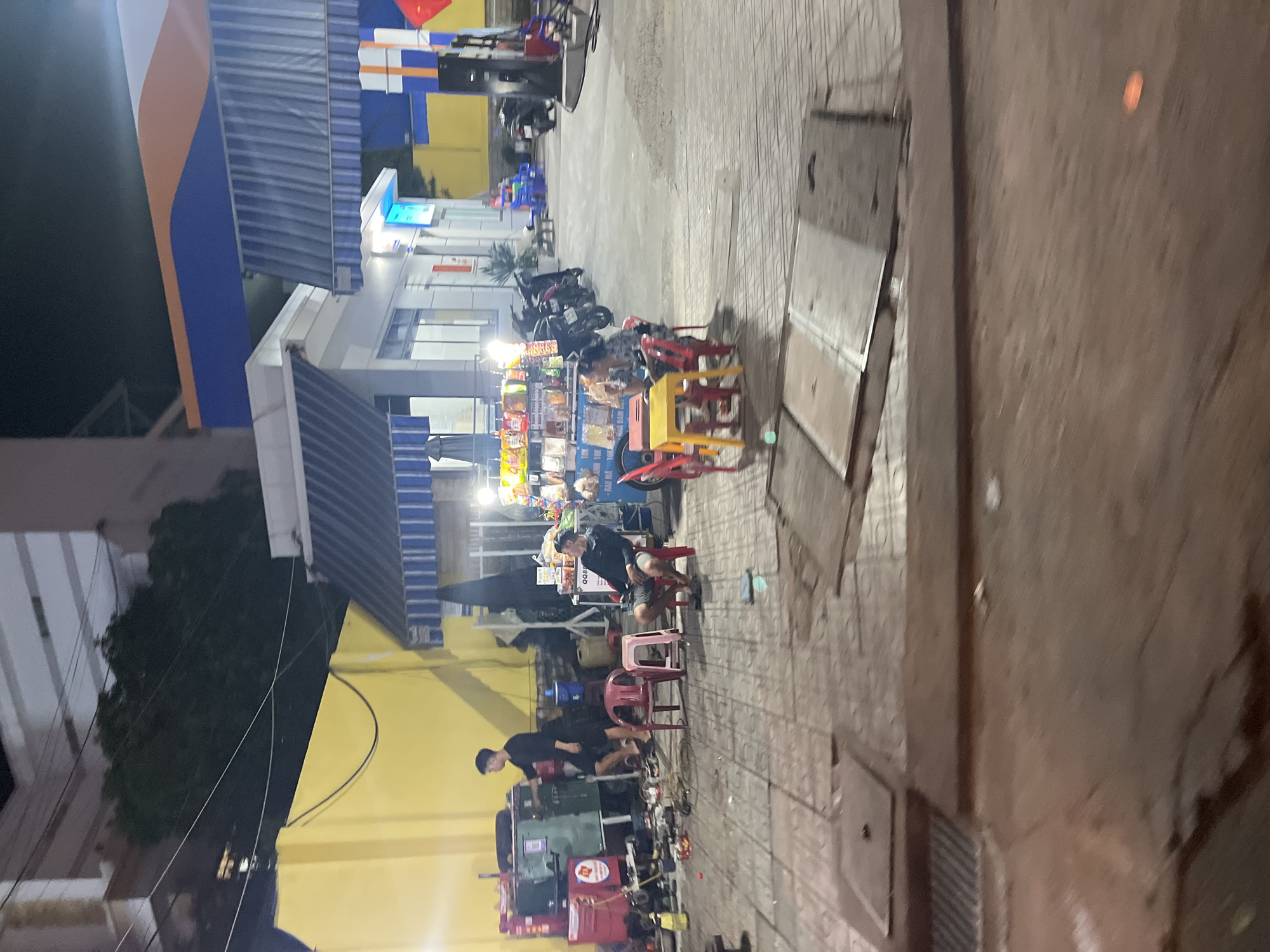 The image depicts a street scene in Bac Nha Trang Ward, Vietnam. The scene includes a building with blue and yellow architectural elements, possibly a gas station. Various parked motorcycles are visible near the building. In the foreground, there's a mobile food stall with food items on display and people gathered nearby. One individual is seated on a stool, possibly a customer. Another person is standing near the stall. Various mechanical devices and equipment are on the ground beside the stall. The street is paved. The overall lighting suggests it is either early evening or nighttime.