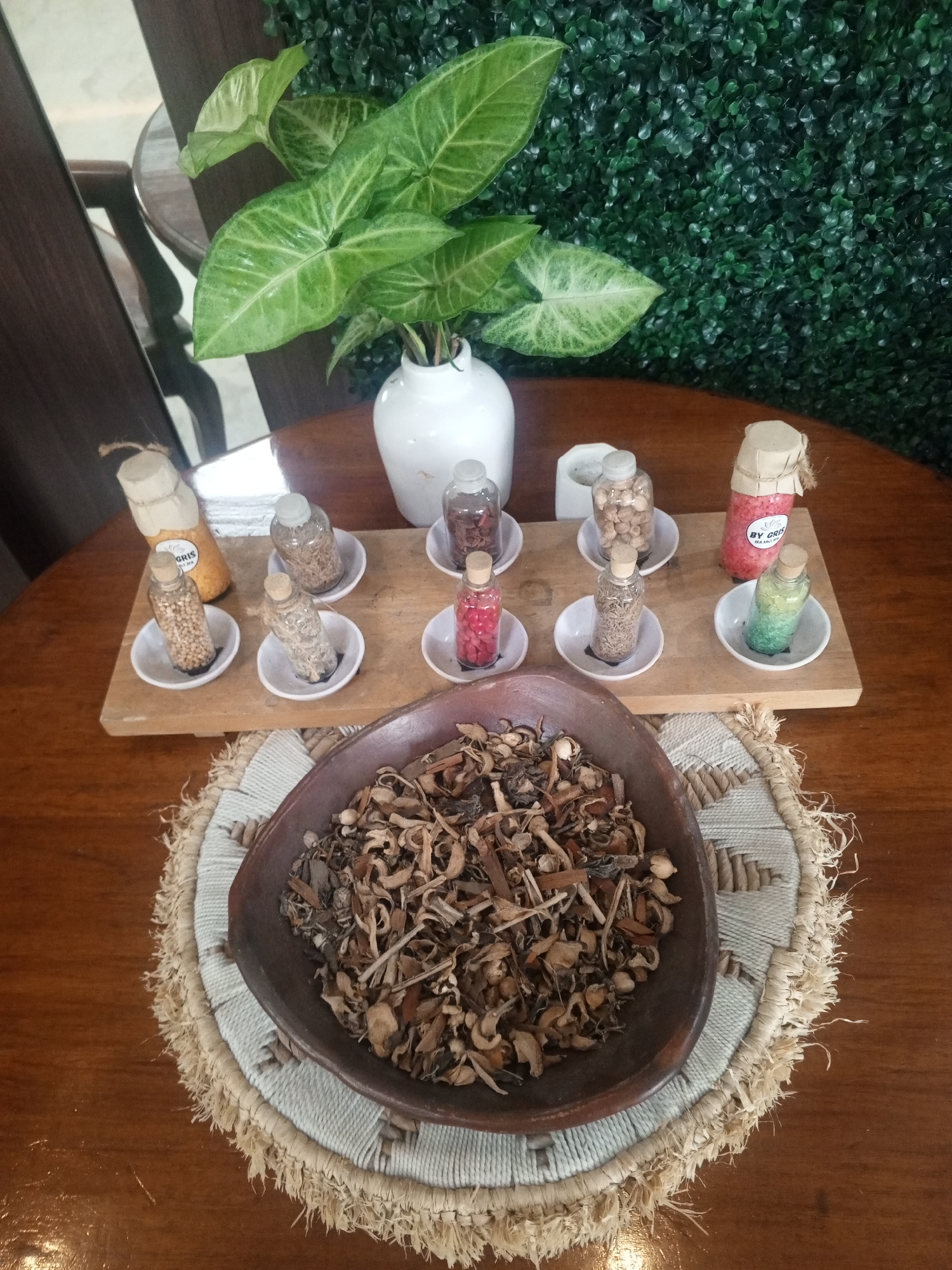 This is a high-angle, medium shot of an arrangement of natural materials on a wooden table. In the background, a wall covered in artificial green foliage serves as a backdrop. A white vase containing a lush green plant with large, heart-shaped leaves is positioned to the left. In the foreground, a dark brown, shallow bowl is filled with an assortment of dried, natural elements that appear to be herbs, roots, and possibly small seed pods, suggesting an apothecary or natural product display. This bowl rests on a woven mat with a fringed edge. In the mid-ground, a light-colored wooden board displays several small glass bottles, each containing different dried botanicals or colored granular substances, arranged on small white saucers. One bottle, labeled "BY CRIS," contains pink granules, and another holds green and yellow layered granules. The overall scene suggests a curated presentation of natural ingredients, possibly for sale or display at a spa, herbal shop, or wellness center in Bandung City, Indonesia.