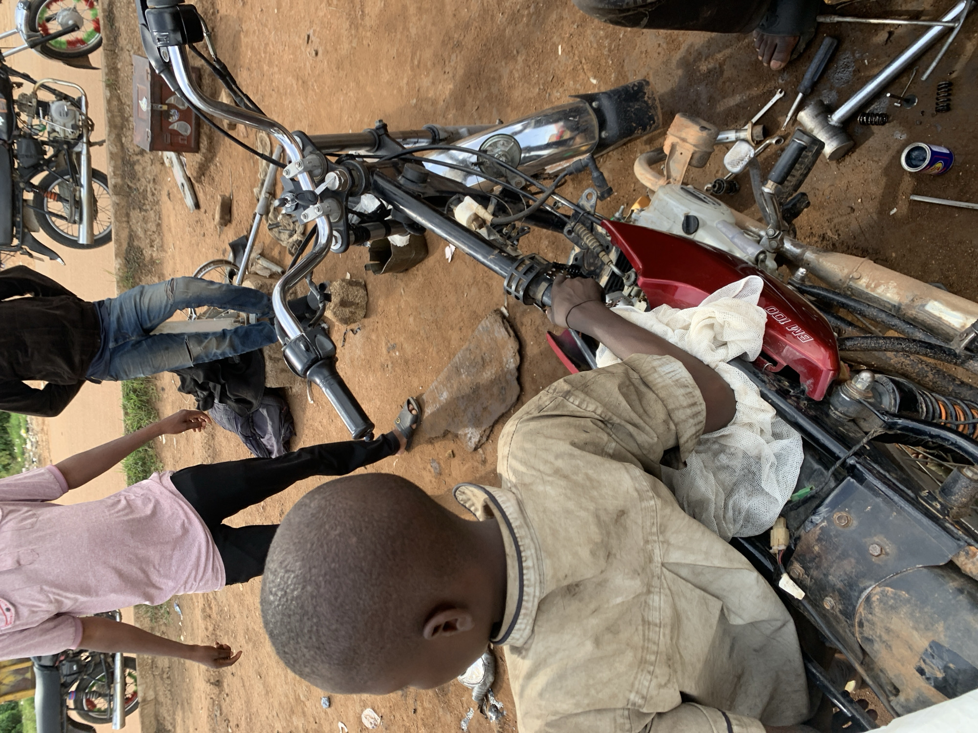 In Barakin Lerawi, Nigeria, an outdoor scene depicts a young boy intently focused on a partially disassembled motorcycle. He is dressed in a dirt-stained tan shirt and appears to be holding a white cloth. His head and shoulders are prominently featured from behind. The motorcycle, a red and black model, is on its side, with various parts and tools scattered on the sandy ground around it. In the background, two men are visible. One, wearing a pink polo shirt and black pants, stands near another motorcycle. The second man, in ripped blue jeans and a dark jacket, stands further back. The environment suggests a working area, possibly a mechanic's shop or roadside repair spot, characterized by loose soil, rocks, and other debris.
