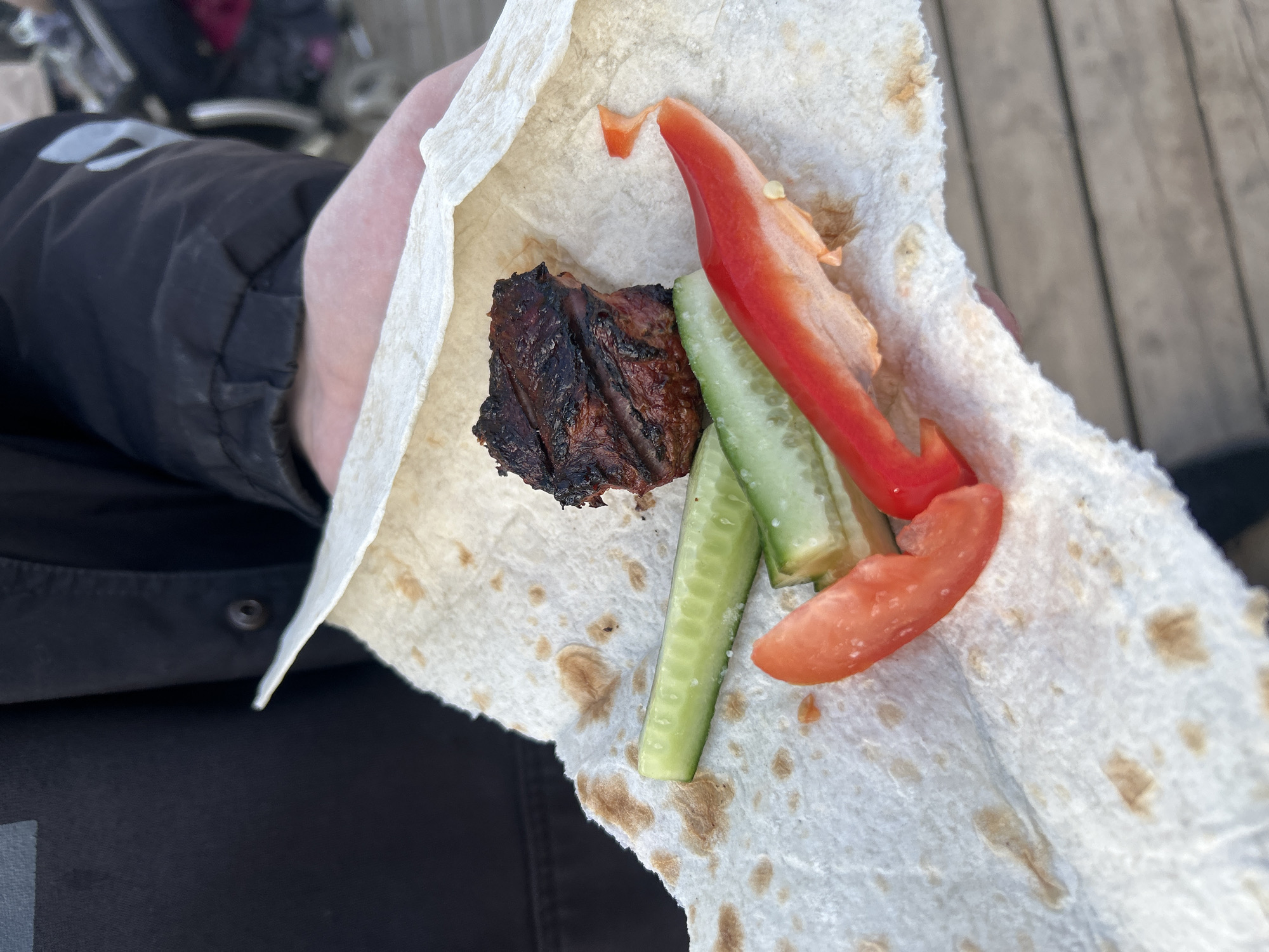 A person holds a flatbread wrap in their hand, containing grilled meat, sliced cucumbers, and sliced red peppers. The location appears to be outdoors in Chaikovsky, Russia. The background includes a blurred view of wooden planks and other individuals, suggesting a casual outdoor setting. The focus is on the open wrap, presenting the contents clearly. The person's dark-colored jacket and parts of their pants are also visible in the frame.