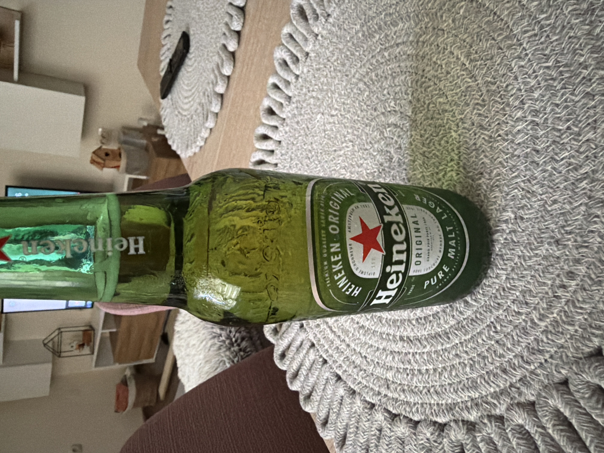 A green glass bottle of Heineken beer is placed on a round, grey woven placemat on a wooden table. The bottle is partially full and condensation is visible on its surface. The label on the bottle clearly displays the Heineken logo, including the red star and the brand name. In the background, a television screen is visible, along with some shelves and decorative items, suggesting a living room or dining area. The overall scene is indoors, with soft lighting. The image does not provide any information about the city of Pazardzhik, Bulgaria.