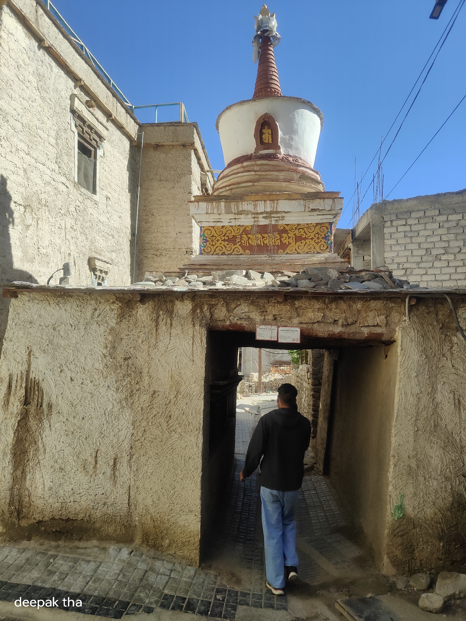 A full shot captures a man in a black hoodie and blue jeans walking away from the camera into a narrow alleyway. The alleyway's walls are textured with a rough, light-colored plaster and the ground is paved with dark and light rectangular tiles. The man is positioned centrally in the frame, with his back to the viewer. Above and behind the alleyway, a white stupa with a red and gold decorative band is visible against a clear blue sky. The stupa is adorned with a gilded emblem in a niche. To the left of the stupa, a rough-textured, light-colored building with a small, dark window is visible. To the right, a partially constructed building with white brickwork and exposed rebar rises. The scene appears to be within the city of Leh, India, as indicated by the architectural style and the watermarked text "deepak tha" in the lower left corner.