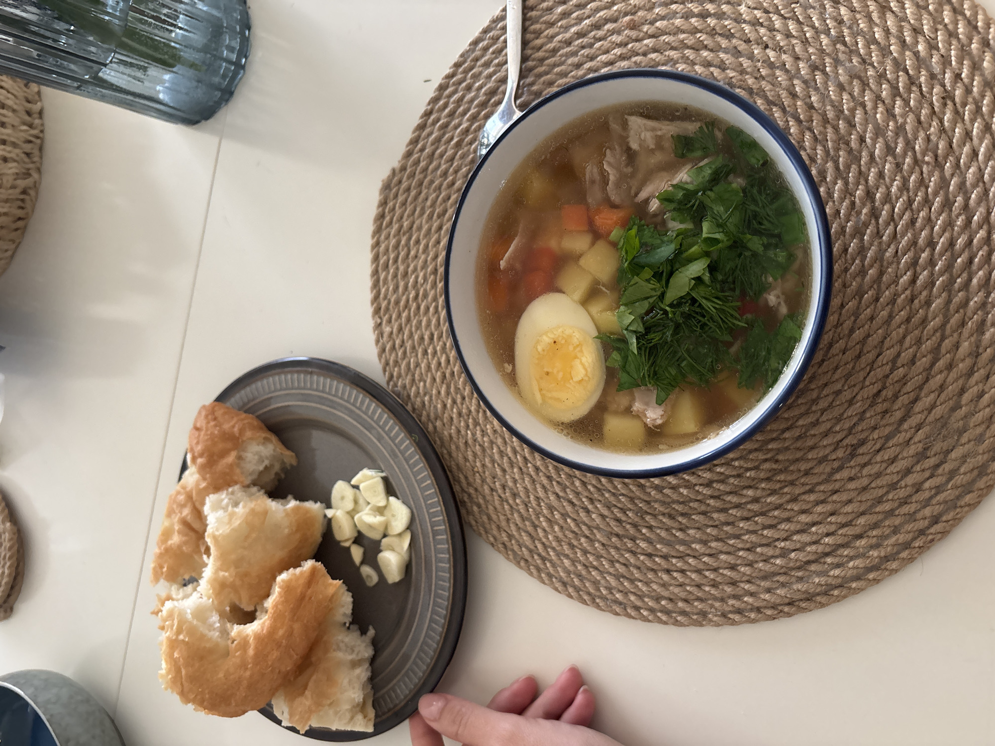 A high-angle shot captures a meal arrangement. A bowl of soup with a halved boiled egg, greens, and vegetables rests on a round, braided mat. Next to it, a gray plate holds bread pieces and sliced garlic. A person's hand appears near the plate. Utensils are present. A blue glass object is partially visible in the upper left. The scene takes place in Krasnoyarsk, Russia.