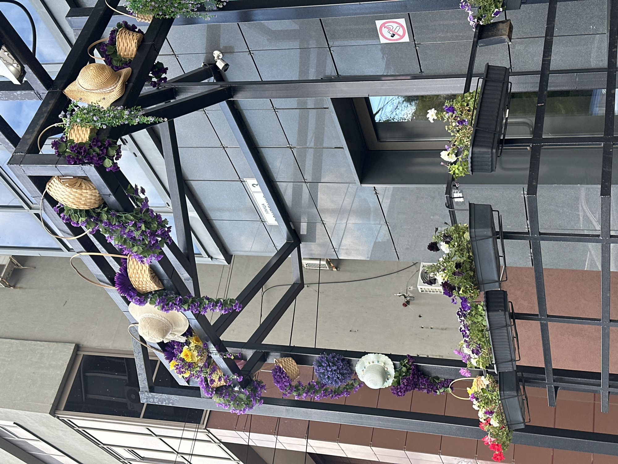 A black metal structure is decorated with hanging baskets of purple and yellow flowers, as well as straw hats. The structure is attached to a modern building with a gray tiled facade. In the foreground, there are three black planters filled with various flowers, including red, pink, purple, and white petunias. The planters are mounted on a black metal railing. Above the planters, a white hat with a ribbon is hanging from the metal structure. On the building's facade, a security camera and a "No Smoking" sign are visible. The background shows another building with a gray concrete exterior and tinted windows. The overall scene appears to be an outdoor decoration in a city environment.