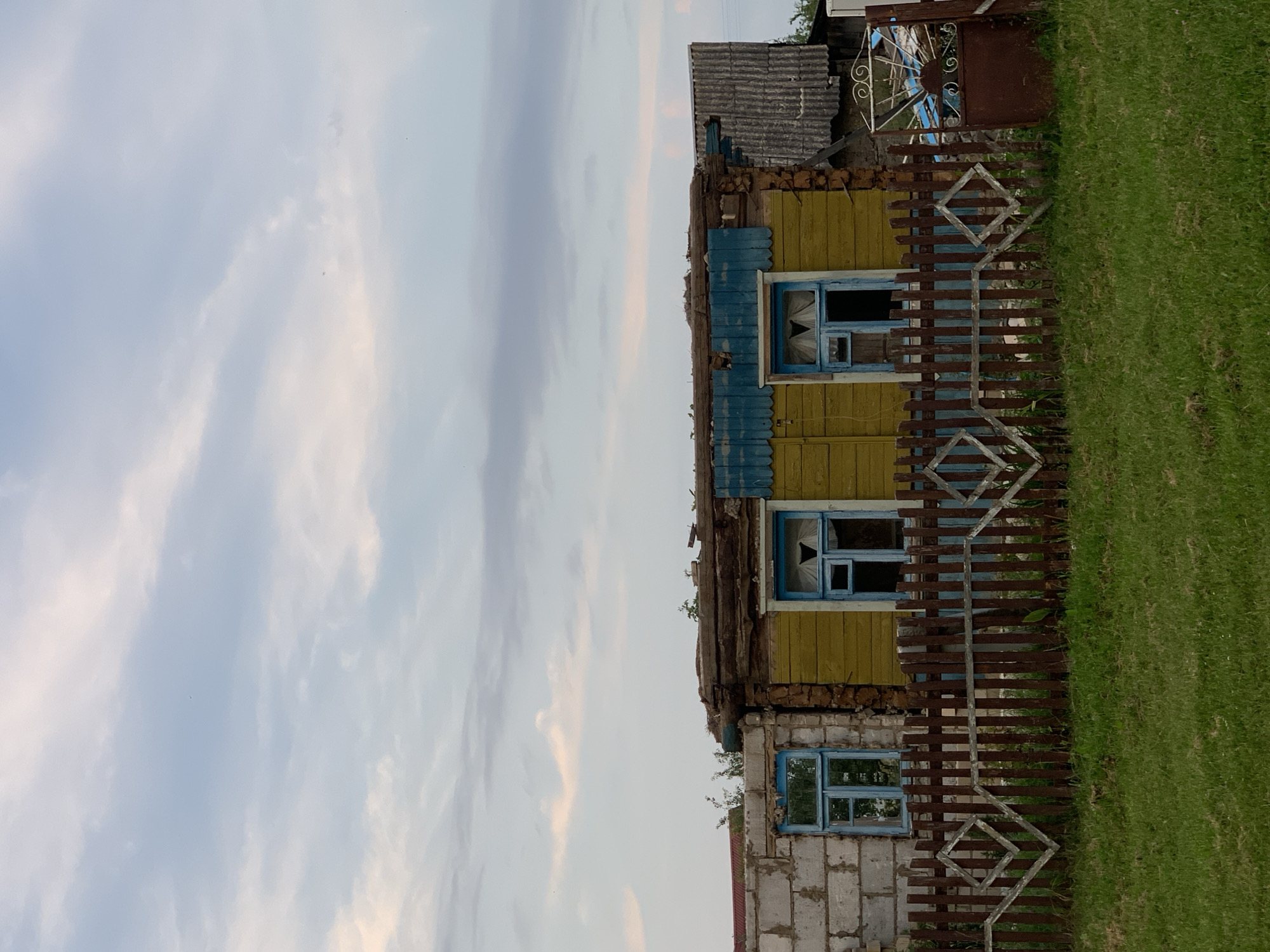 This image depicts the exterior of a dilapidated wooden house located in Kapyĺ, Belarus. The house exhibits signs of disrepair, with a partially collapsed roof and weathered wooden siding painted yellow. Two windows with blue frames are visible on the front of the house, one made of cinder blocks and the other of wood. A brown wooden fence, decorated with diamond-shaped patterns, runs in front of the house. The scene is set against a cloudy sky, and a green grassy area is in the foreground. The overall impression is one of abandonment and decay.