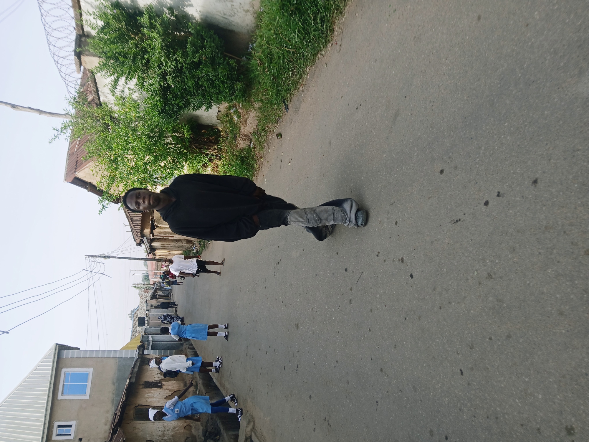 The image displays a street scene captured in daylight, appearing to be in a residential area. The photograph is oriented vertically (portrait mode), but presented rotated 90 degrees clockwise.
In the foreground, a young man, appearing to be the primary subject, stands on the right side of an asphalt road. He is dark-skinned, wearing a black long-sleeved top, faded grey jeans, and dark shoes. He looks directly at the camera with a neutral expression, with his left foot slightly raised as if mid-step. Behind him and to his right, green bushes and foliage grow alongside a wall topped with barbed wire.
The road extends from the bottom of the frame towards the top distance. Along the left side of the street, several children, likely school-aged, are visible. They are dressed in light blue and white school uniforms, including dresses or skirts, white tops, and for some, white head coverings. They are walking or standing, with their backs mostly turned to the camera. Further down the street on the left, an adult male in a white t-shirt and dark shorts is also present.
Buildings line both sides of the street. These include a mix of single and two-story structures with varying architectural styles; some have corrugated metal roofs, while others feature painted concrete walls. On the far left, a light-colored building stands out with a modern white-framed window featuring a blue-tinted pane. Utility poles with numerous overhead wires crisscross the bright, clear sky in the upper middle of the frame.
The overall activity depicted is daily life on a street, with people moving about. The presence of children in school uniforms suggests it is a weekday during school hours. The weather appears sunny and clear. No visible text is present in the image.