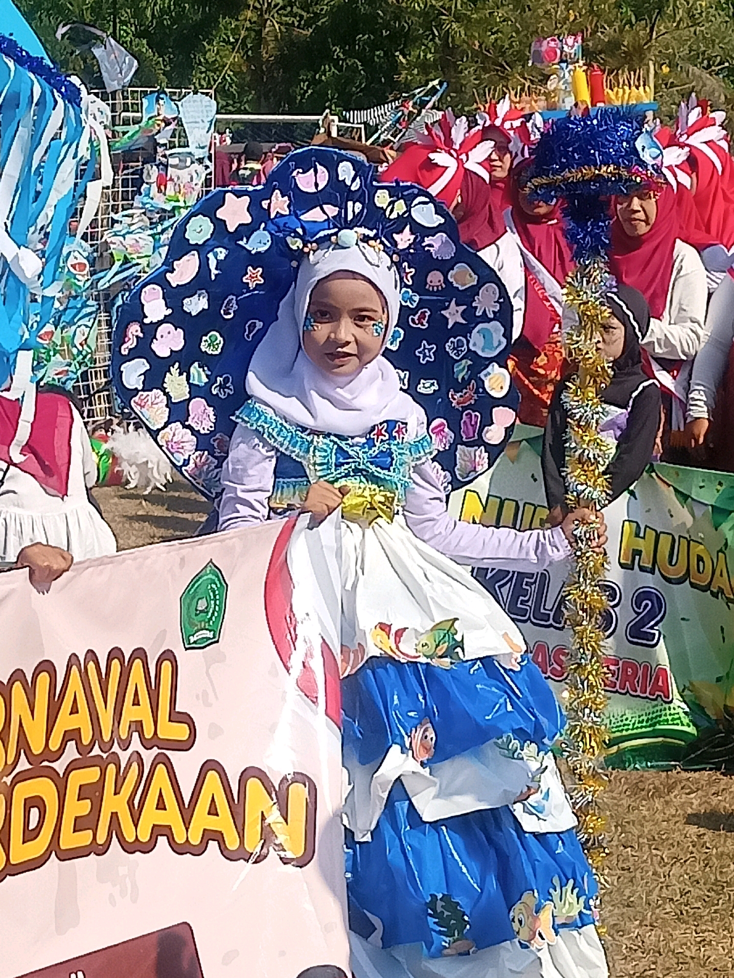 In an outdoor daytime scene in Sumberlawang, Indonesia, a young girl wearing a white hijab and a vibrant, ocean-themed costume stands center frame, holding a banner. Her costume features a large, blue, shell-shaped headdress adorned with various sea creatures like fish, octopuses, and starfish. She wears a white shirt with blue and gold trim and a layered blue and white skirt decorated with more aquatic figures. Glitter is applied to her cheeks, and she holds the banner with both hands. The banner, mostly pink and white, displays yellow text that reads "CARNAVAL MERDEKAAN". Behind her, other participants in similar colorful costumes, some with red and white headwear, are visible, suggesting a parade or festival. A white wire fence and green foliage form the background. The ground is covered in dry grass. Another banner to the right displays text that partially reads "HUDAN KELAS 2".