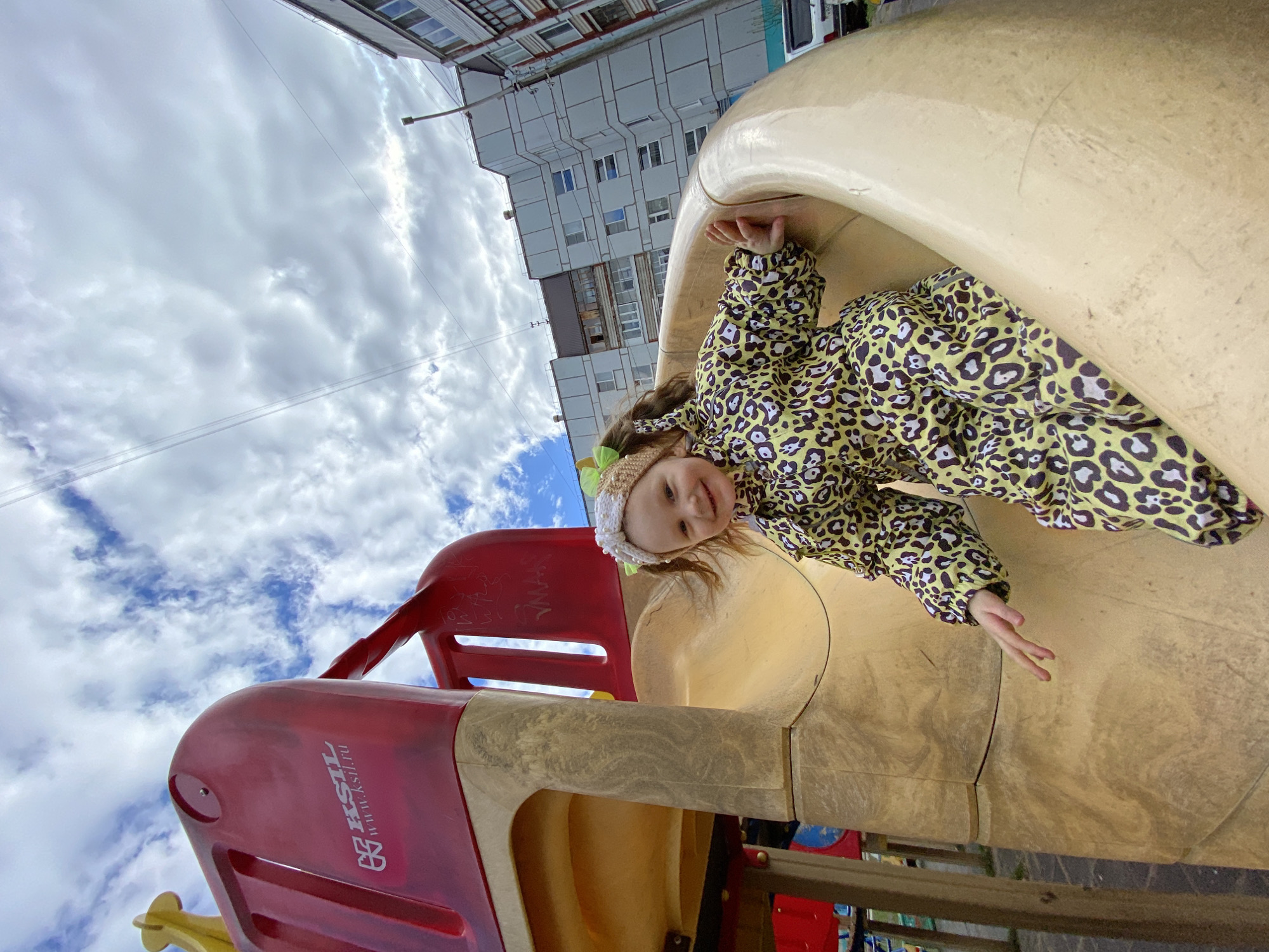 A young child in a yellow and black leopard-print snowsuit is playing on a red and beige playground slide. The child is wearing a knitted hat. The playground appears to be in an urban setting, possibly Chaikovsky, Russia, given the presence of the website "www.ksil.ru" on the slide, which refers to a Russian playground equipment manufacturer. The sky is cloudy. A multi-story building is visible in the background.