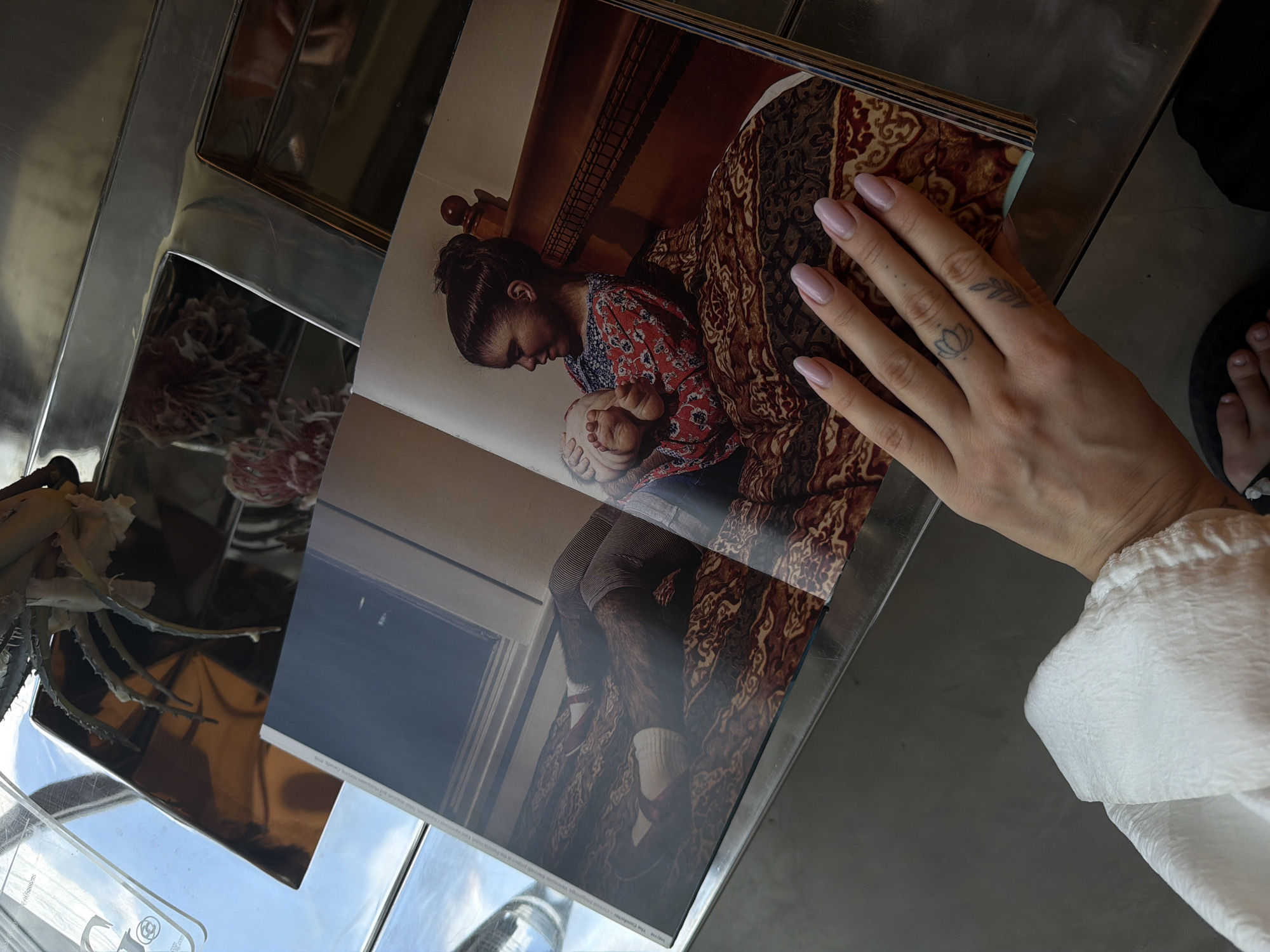 An open magazine is positioned on a metallic surface. The magazine displays an image of a young girl holding a baby on a bed. To the right of the magazine, a person's hand with light pink fingernails and subtle tattoos on their fingers is visible, resting on the magazine. A portion of a white fabric is also visible in the lower left corner of the image, and a pair of feet are seen at the bottom. The environment suggests an indoor setting, potentially a home or a studio, with natural light filtering in from a window, creating reflections on the metallic surface. There is no specific indication of the location being in North Kuta, Indonesia.