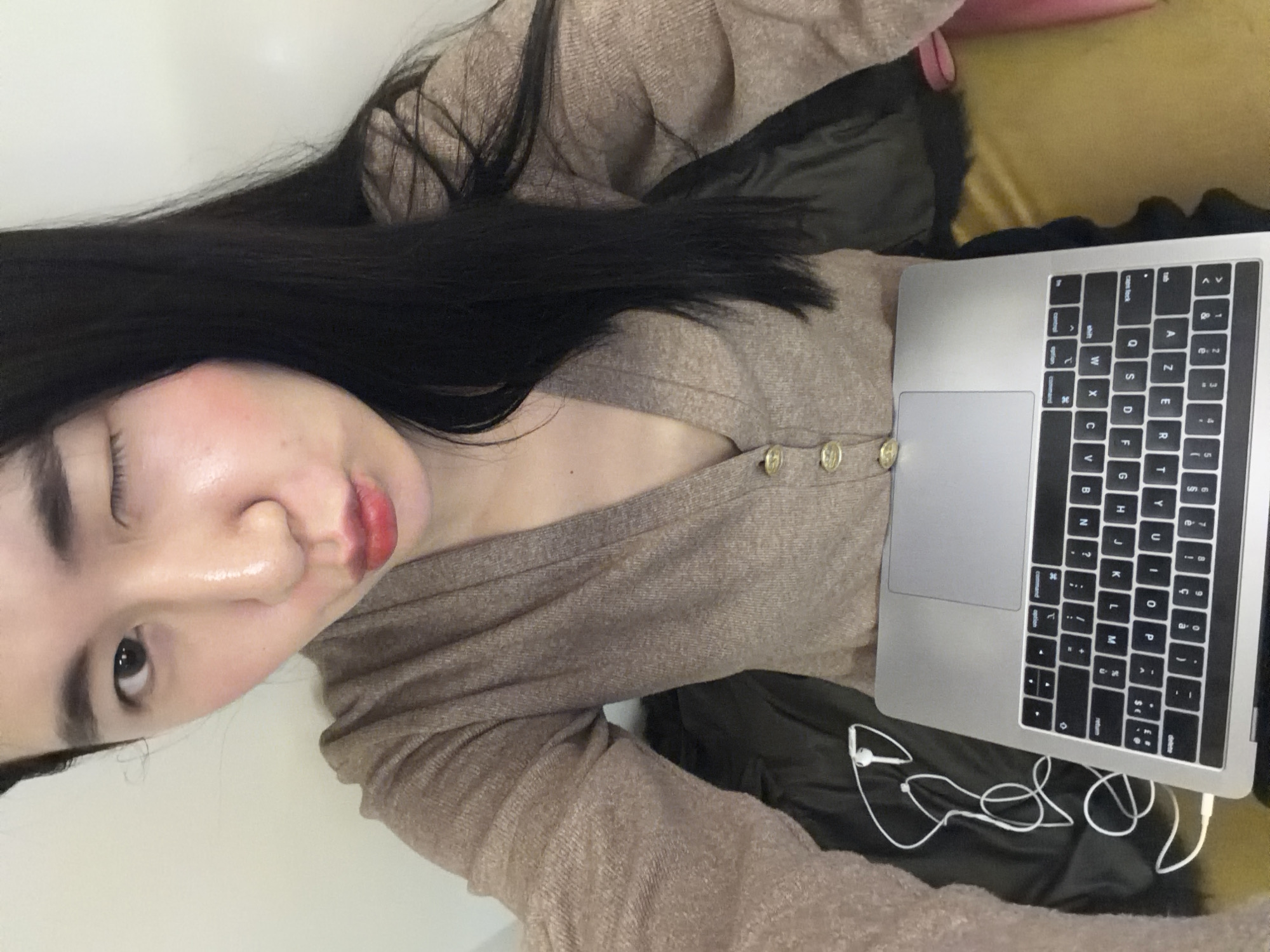 The image shows a person with long, dark hair wearing a light brown cardigan with three buttons, lying on what appears to be a dark fabric-covered surface, possibly a beanbag or cushion. A silver laptop is positioned on the person's lap, with a white pair of wired earphones tangled near the laptop's edge. The person has one eye closed and is looking towards the camera. The background is out of focus. There is no visual information in the image to determine if the photo was taken in Paris, France.