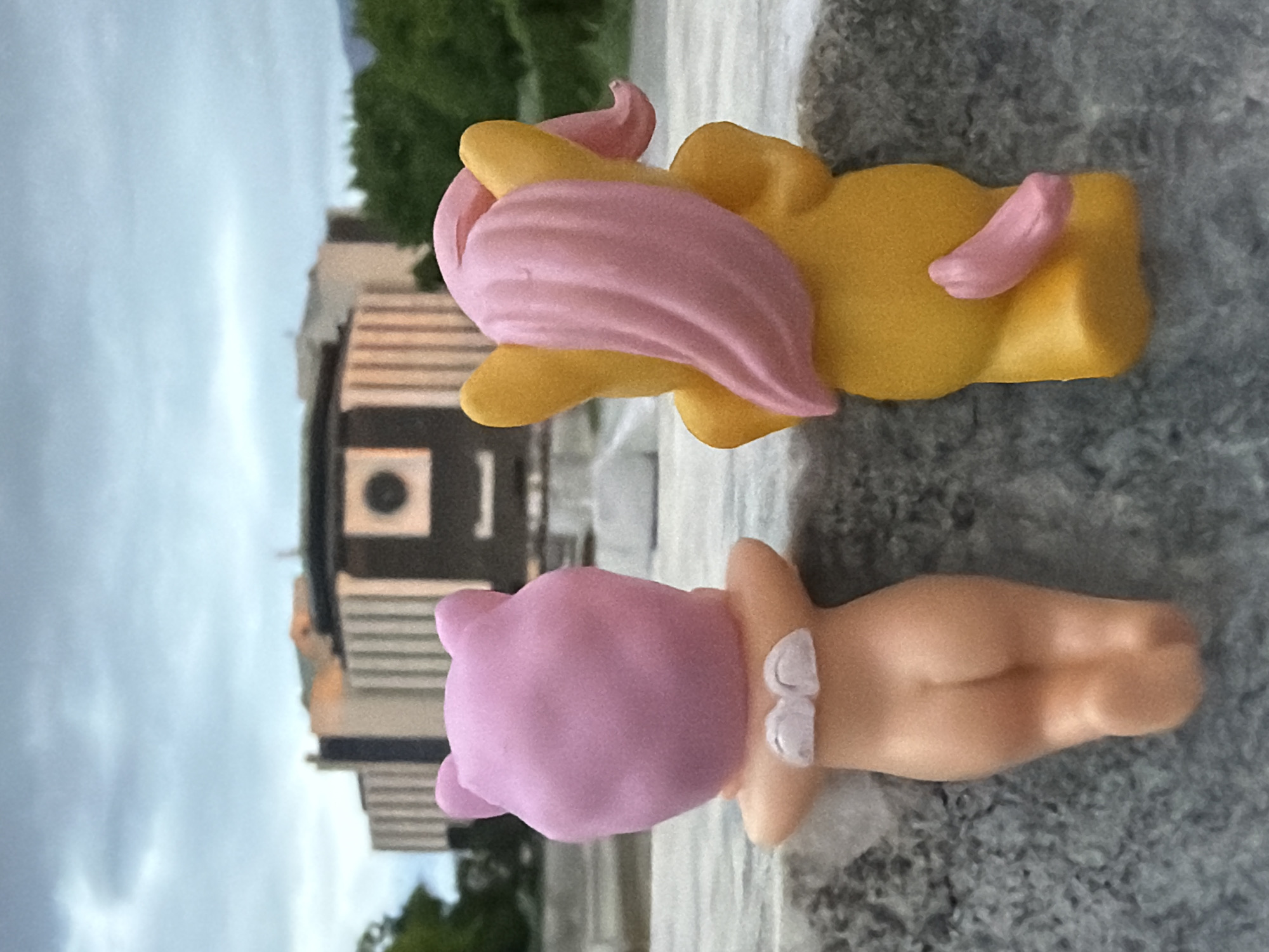 Two plastic toy figures are arranged on a textured, grey surface that appears to be concrete. The top figure is yellow with a pink mane and tail. The bottom figure is pink-headed with a fleshy body and a white bow on its back. In the background, a building with dark and light horizontal stripes can be seen, along with green trees and an overcast sky. Based on the architectural style of the building, the location is likely Sofia, Bulgaria.