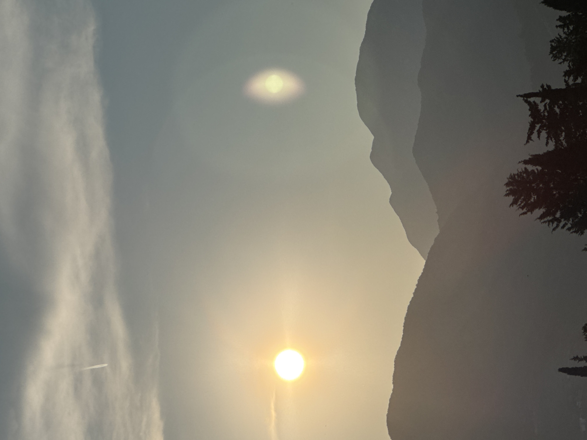 This image captures a mountain landscape at sunrise or sunset. The sun, a bright orb of light, is visible in the sky, casting a warm glow. There are mountains in the background, silhouetted against the sky. On the left side of the image, there are wispy clouds and a contrail from an airplane, indicating that the photo was taken during the day, likely morning or late afternoon. The overall mood of the image is serene and peaceful. The lighting suggests it is either early morning or late evening. There is no visible text in the image.