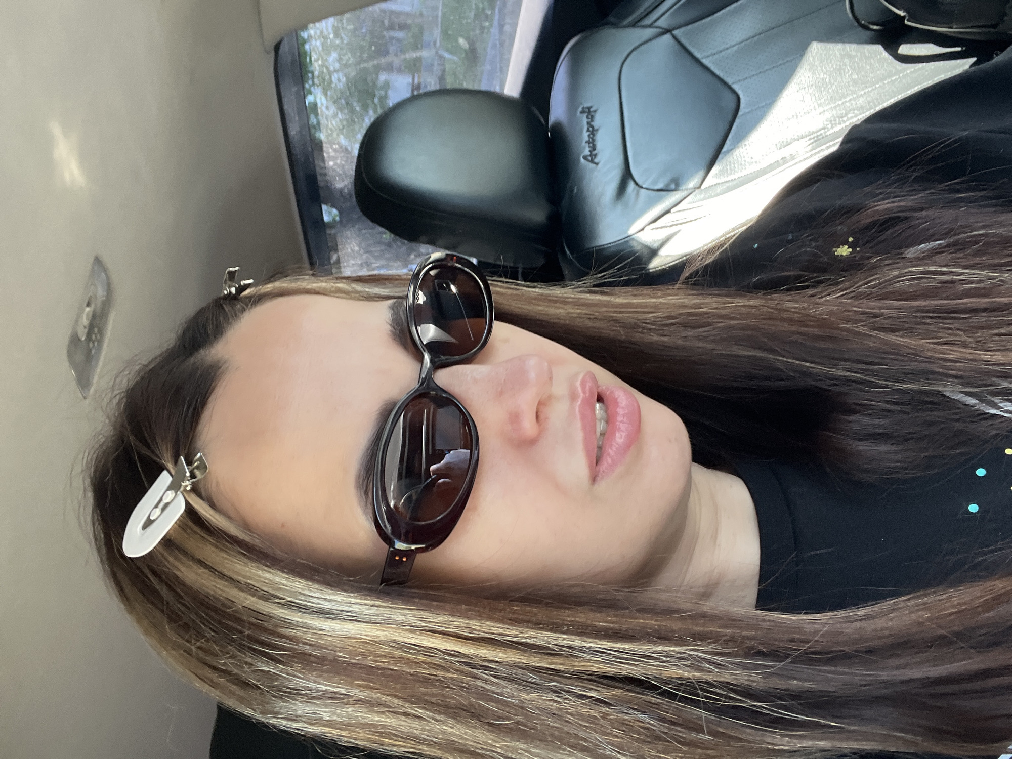 A woman is sitting in a vehicle, possibly a car, with a black seat visible in the background. She is wearing sunglasses perched on her nose, and her hair is styled with highlights. There are white hair clips in her hair near her forehead. She is wearing a black shirt with small, colorful polka dots on it. The vehicle's window is visible, showing trees and outdoor scenery.