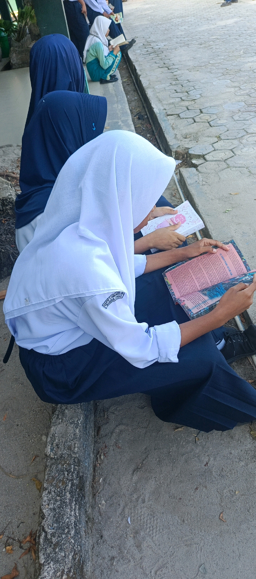 In the village of Desa Sarang Mandi, Indonesia, two young Indonesian females are seated on a concrete ledge. One female, in the foreground, wears a white hijab and a white school uniform shirt with dark pants. She is engrossed in reading an open book with pink pages and illustrated borders. To her left and slightly behind, another female also wears a dark blue hijab and a similar school uniform, but she is partially obscured. In the background, another student in a light green uniform is visible sitting and reading a book. The environment appears to be an outdoor area with a textured, paved walkway to the right and sandy ground with some scattered leaves to the left. The overall scene suggests a moment of focused study or leisure reading among students.