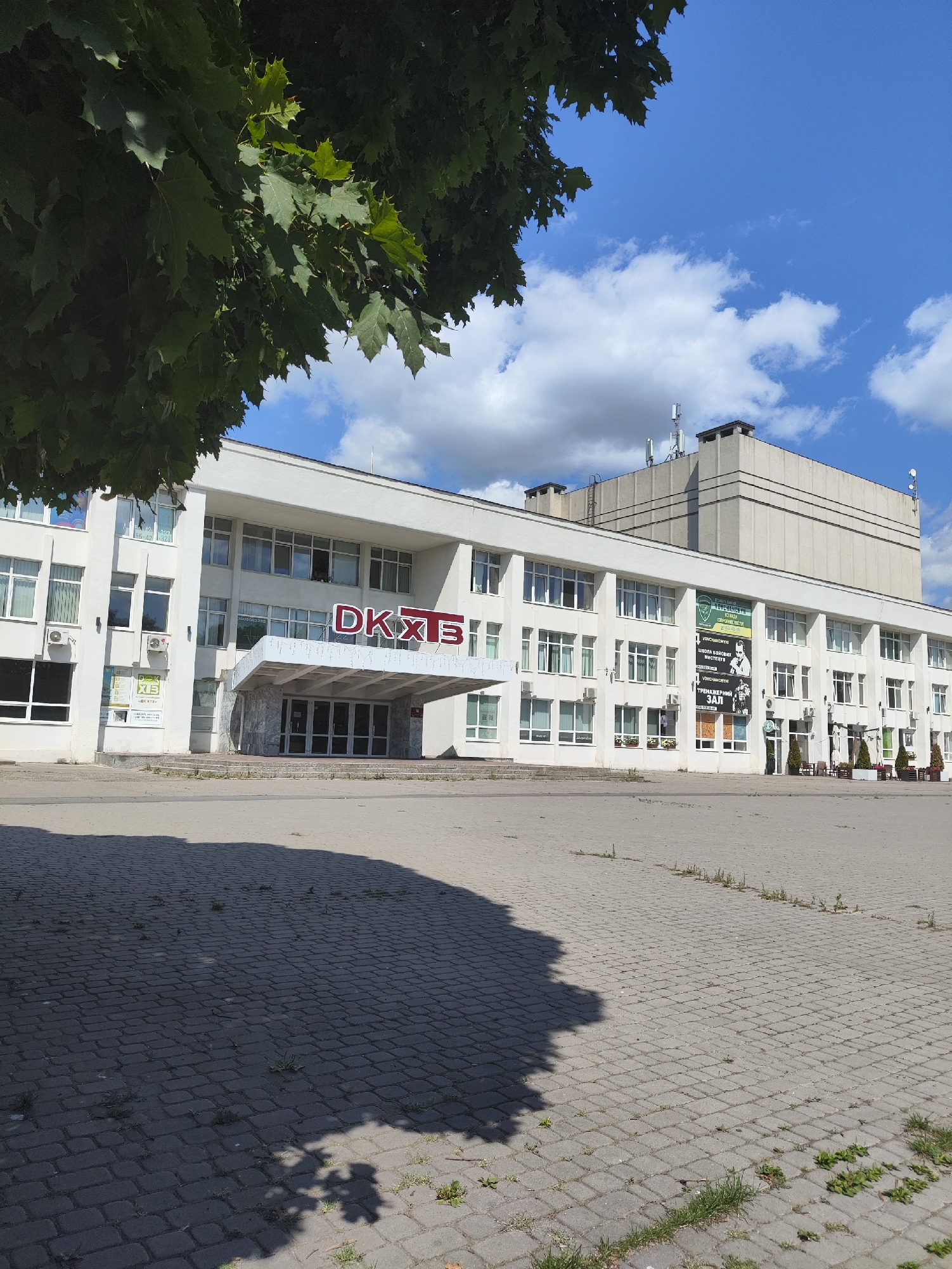 This is a full, eye-level, exterior, medium shot of the "DKKhTE" building in Kharkiv, Ukraine, on a sunny day with scattered clouds. The building is white with a prominent marquee entrance. The letters "DKKhTE" are displayed in red above the entrance. A large tree with dark green leaves partially obscures the upper left corner of the image. The foreground consists of a paved plaza area with a shadow cast from the tree. The building has many windows, and several posters are visible on its facade. The sky is blue with white, fluffy clouds.
