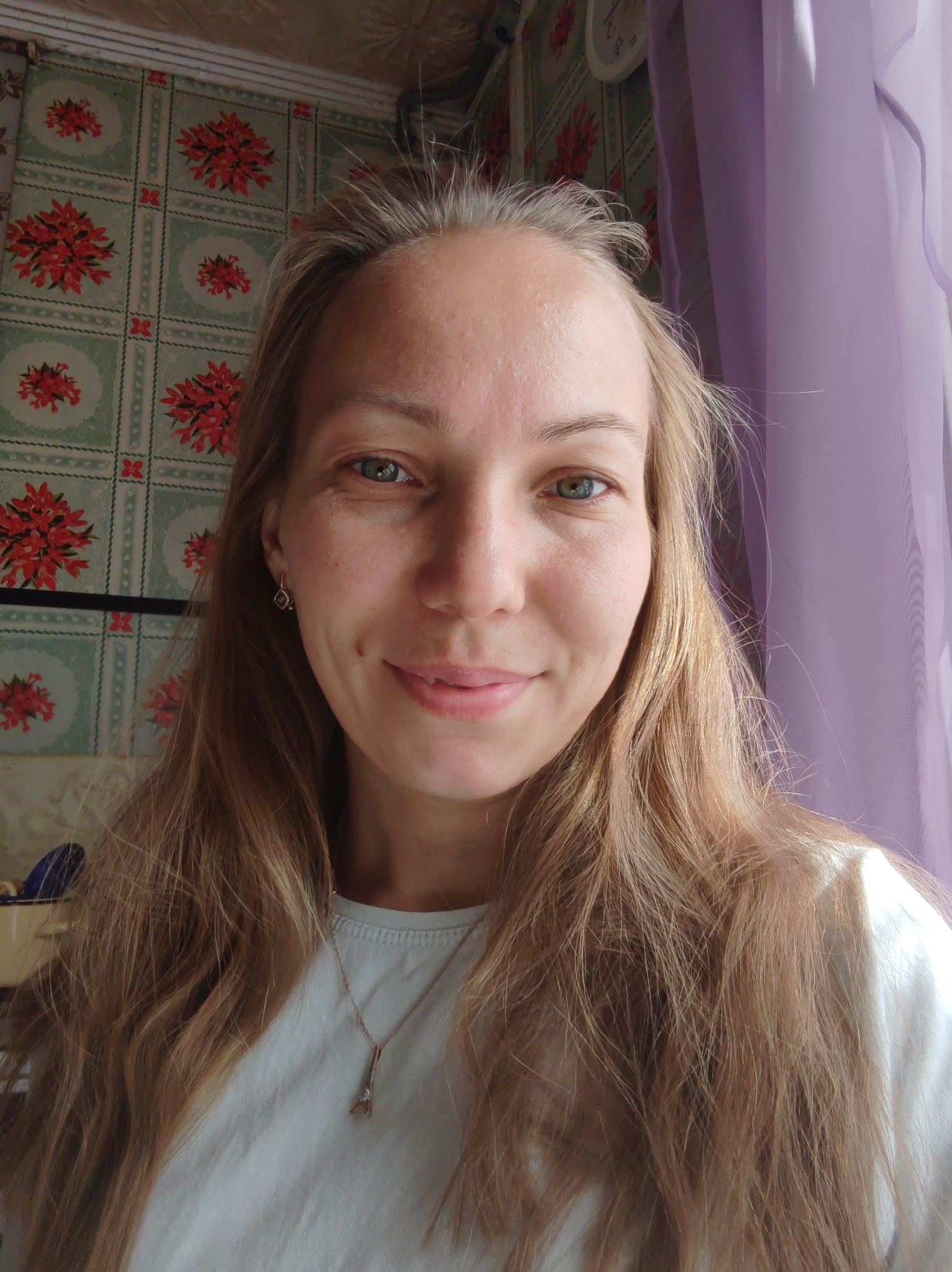 A young woman with long, wavy blonde hair and light green eyes is taking a selfie indoors. She is wearing a light blue t-shirt and a silver pendant necklace shaped like the Eiffel Tower. A small, diamond-shaped earring is visible in her left earlobe. Her skin is fair with some freckles and a dimple on her right cheek. She is smiling gently. Behind her, a wall is decorated with a pale green wallpaper featuring a repeating pattern of red floral arrangements. To her right, a sheer purple curtain is partially visible, suggesting the presence of a window. The lighting in the room appears to be natural, coming from the direction of the window. The background also shows a glimpse of what might be a clock on the wall. The overall scene suggests a casual, everyday setting. The image is a medium shot, focusing on the woman's face and upper body.