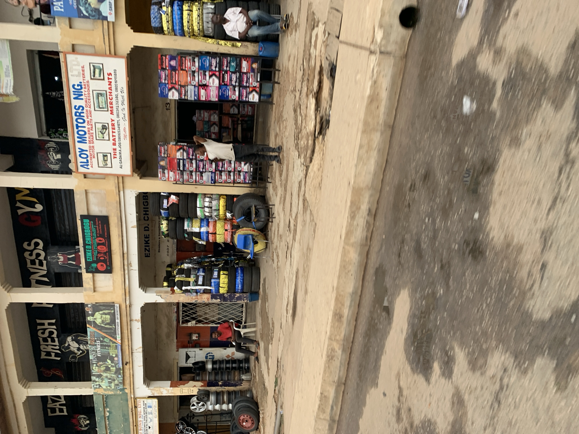 This street-level view in the New Stadium area of Jos, Nigeria, captures a commercial district. The main focus is a row of storefronts. Two businesses, ALOY MOTORS NIG. LTD. and EZIKE D. CHIGBOGU, are prominently featured as battery and tire merchants, their storefronts displaying stacked tires and batteries. Another business called Easy & Faster Fitness is seen on the left. People are present near the shops. The architecture is simple, with painted storefronts and open entrances. The sidewalk is concrete and somewhat worn.