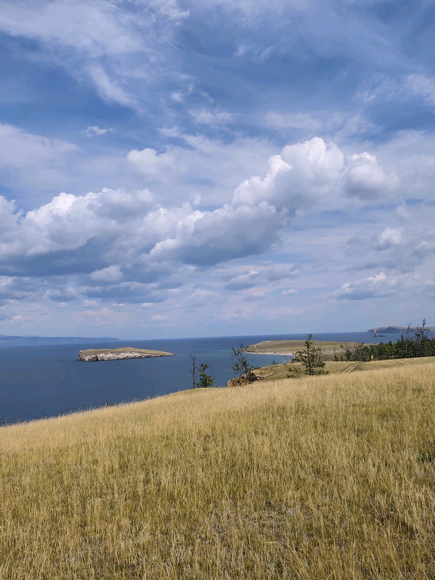 The image captures a scenic view of a large body of water, likely Lake Baikal, near Kharantsy, Russia. The foreground is dominated by a rolling hill covered in dry, golden grass, suggesting a summer or early autumn season. The grassy slope leads down towards the water.
In the middle ground, a small, rocky island with sparse vegetation sits in the calm, deep blue water. Further in the distance, the coastline curves, revealing a sandy beach area with some parked vehicles and scattered trees. More landmass is visible on the horizon, with low-lying hills and a coastline extending to the right. The sky above is a vibrant blue, dotted with large, fluffy white clouds and some thinner, wispy cirrus clouds, indicating a partly cloudy day. A few sparse trees are also visible on the grassy hillsides. The overall impression is one of a natural, expansive landscape.