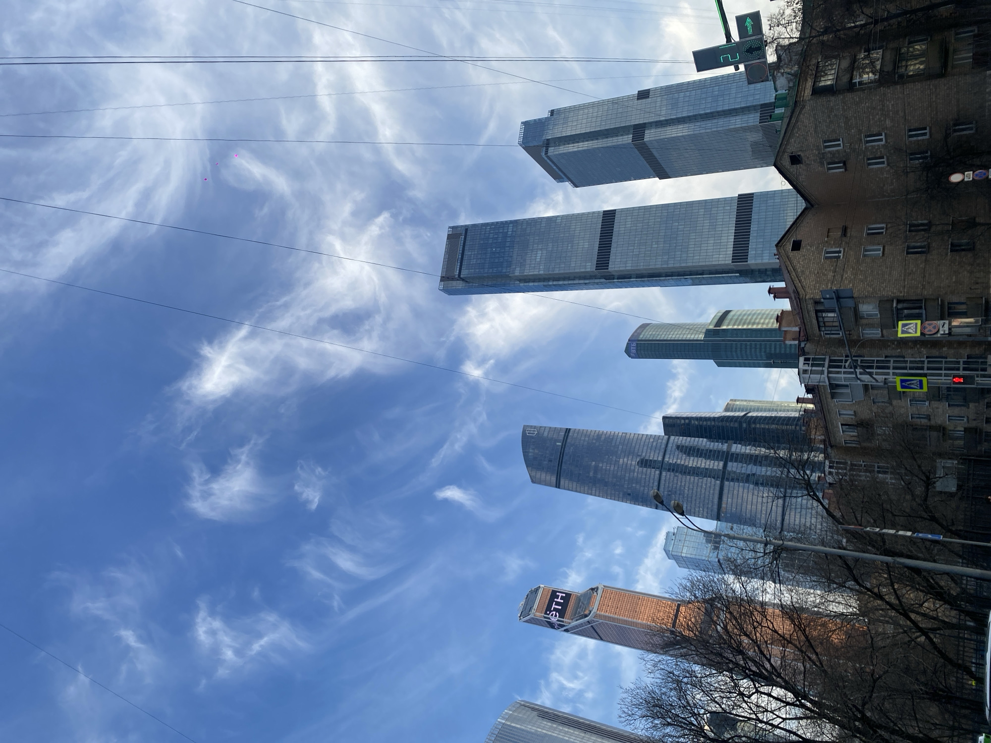 The image depicts an urban landscape under a bright blue sky with wispy white cirrus clouds, crisscrossed by multiple dark overhead wires. Two faint, small magenta blurs are visible high in the sky.
In the mid-ground, a cluster of modern, high-rise glass skyscrapers dominates the background. From left to right, these include: a partially visible tall, light-colored building; a distinct copper-toned angular skyscraper featuring the "ВТБ" (VTB) logo near its apex; a dark-glass tapered skyscraper; and two tall, light-blue glass skyscrapers, one with a horizontal dark band, and the other on the far right also bearing a VTB logo.
In the foreground, below the skyscrapers, are two connected, multi-story brick residential buildings with brown facades and pitched roofs, featuring numerous white-framed windows. Deciduous trees, bare of leaves, line the street in front of and between these older structures and the distant skyscrapers.
Street furniture includes a black lamppost with two luminaires prominent in the mid-ground. Various traffic signs are visible near the older buildings, including a yellow pedestrian crossing sign and a circular 'no stopping' sign. A traffic signal on the far right displays a green arrow pointing right, with the numbers "2" and "4" illuminated. A dark fence runs across the bottom of the frame, with a small portion of a white vehicle roof visible in the bottom-left corner. No identifiable individuals or active movements are discernible.
The scene is set in Khimki, Russia.