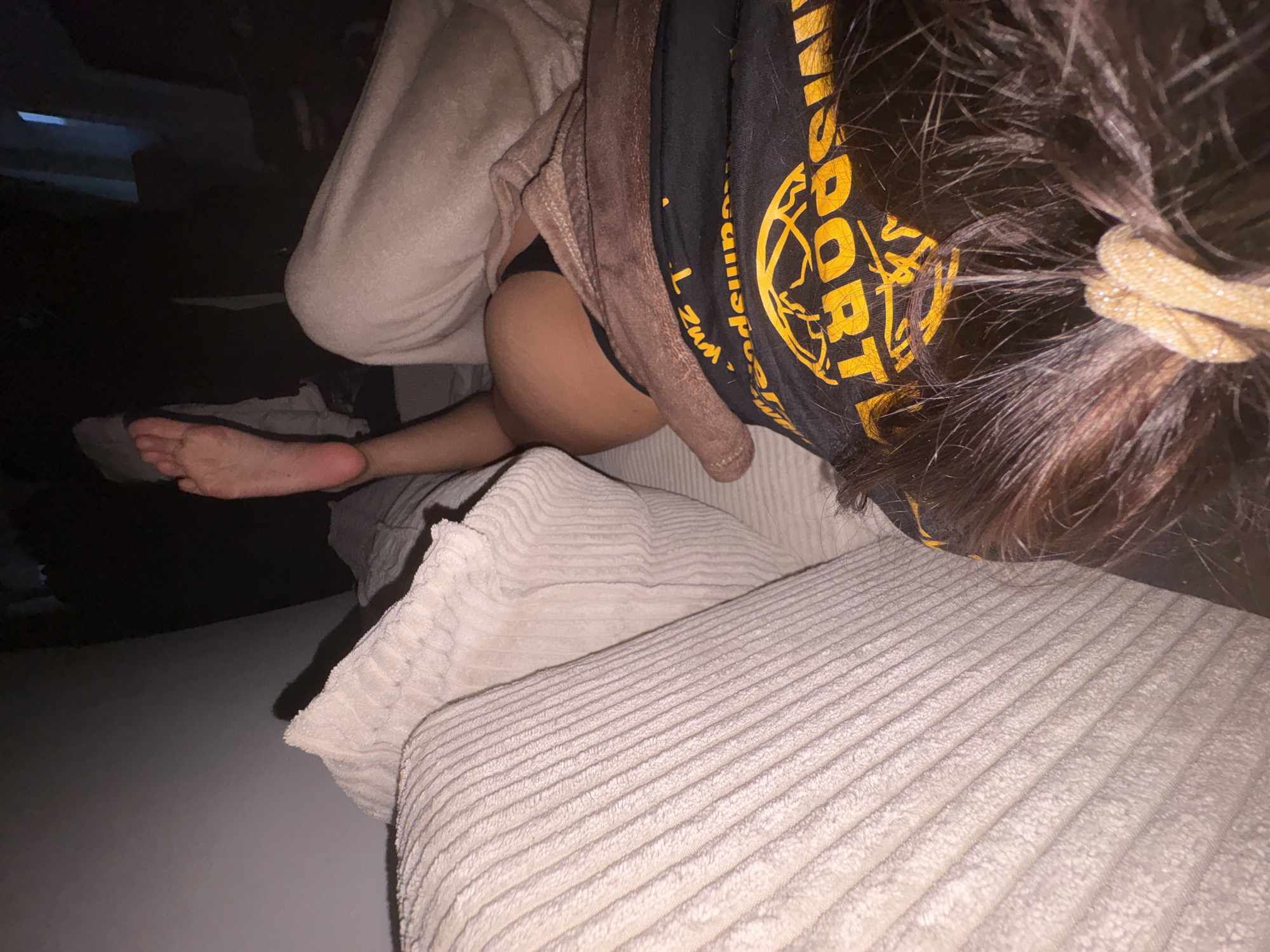 A person is lying on a couch in a dimly lit room, with their right foot propped up on a pillow. The person is wearing a black t-shirt and black underwear. The couch is covered with a light-colored blanket. The room appears to be a bedroom or living room. There are no other people or objects visible in the image. The image does not provide any information about the location in Mönchengladbach, Germany.