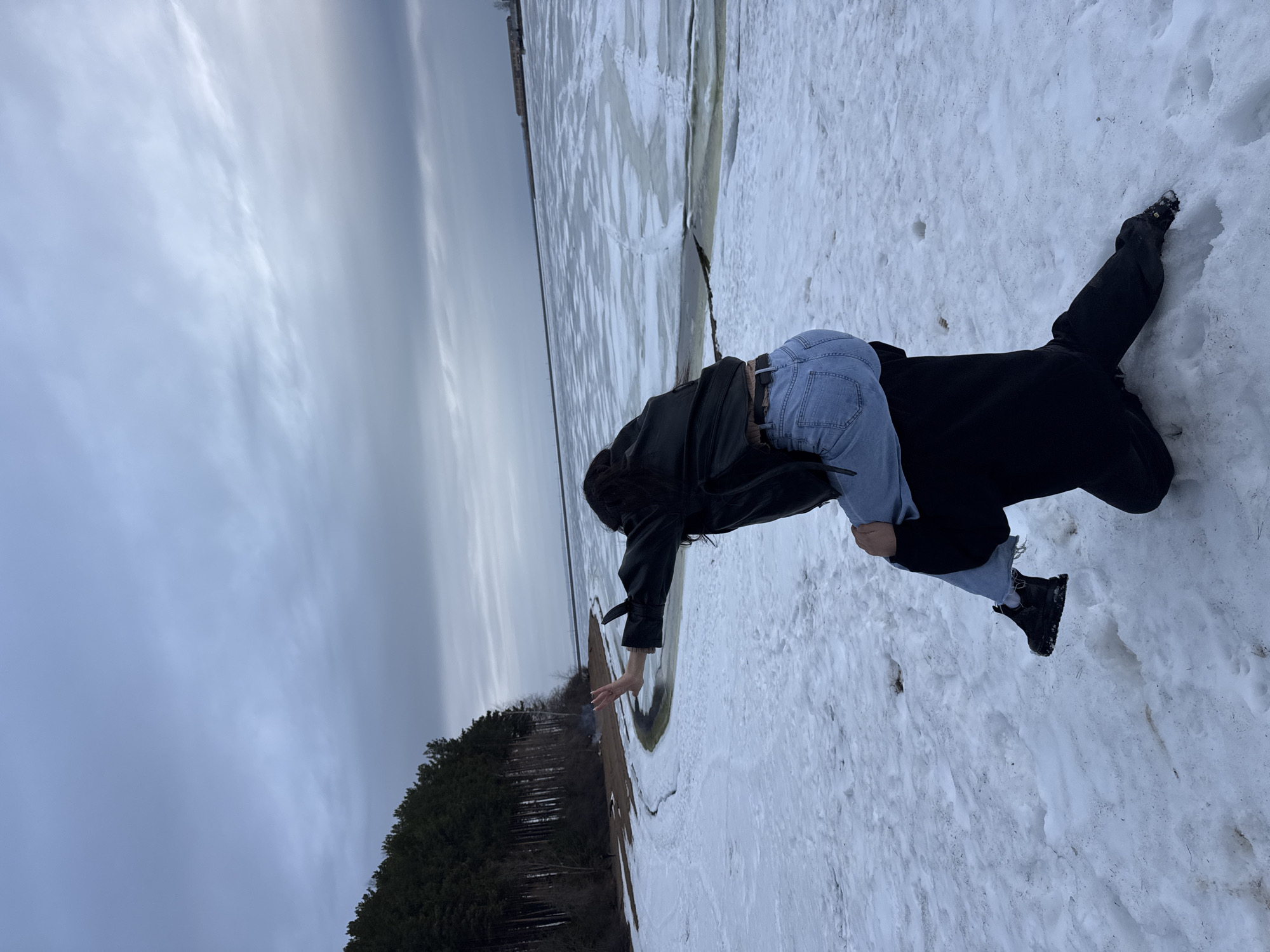 A person in a black coat and blue jeans kneels in the snow, reaching towards a narrow channel of open water separating snow-covered land from a frozen lake. The shoreline is visible with a dense forest on the left side. The sky is overcast. This image was taken in Chaikovsky, Russia.