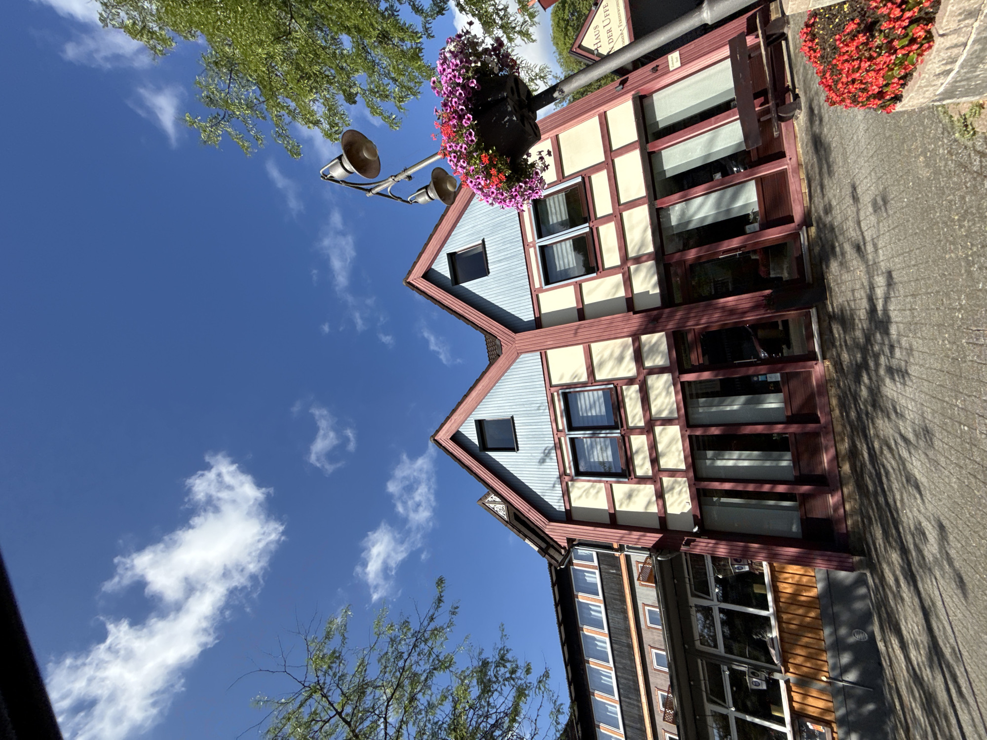 The image shows a street-level view of the front of a building in Bad Sachsa, Germany, with a clear blue sky and white clouds overhead. The building is a multi-story structure with a distinctive architectural style, featuring a combination of light-colored stucco walls with dark red timber framing and light blue siding on the upper gable sections. Large windows and doors with dark red frames are visible on the ground floor. A lamppost with two lamps and a hanging basket filled with colorful flowers stands in front of the building. To the right of the lamppost, a sign is partially visible which reads "Haus Der Uffe" and below that, "Zimmer & Ferienwohnung". A wooden bench is situated in front of the building to the right of the main entrance. Red flowers are also planted in a brick planter in the foreground on the right side of the image. The ground is paved with interlocking bricks and is partially covered in shadows from trees, which are visible on the left and right sides of the frame. The overall scene depicts a bright, sunny day in a European town.