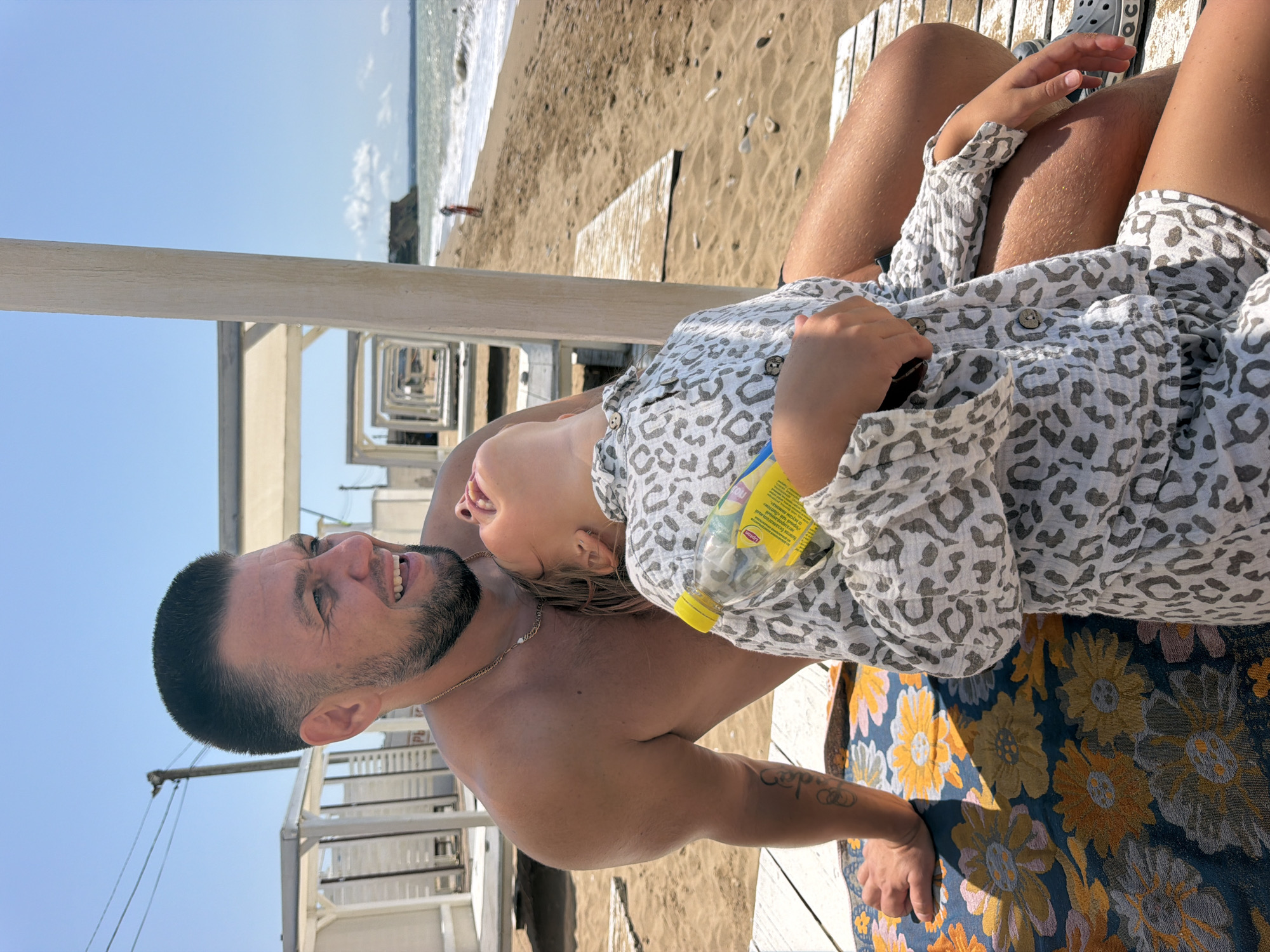 Two people, a man and a child, are sitting on a beach. The man is shirtless and has a beard. The child is wearing a patterned shirt and is holding a bottle of Lipton Iced Tea. They are sitting on a wooden deck with a patterned blanket. The beach has sand and the sea is in the background. There are white cabanas and a cliff in the distance. The location is Качинский округ, Ukraine.