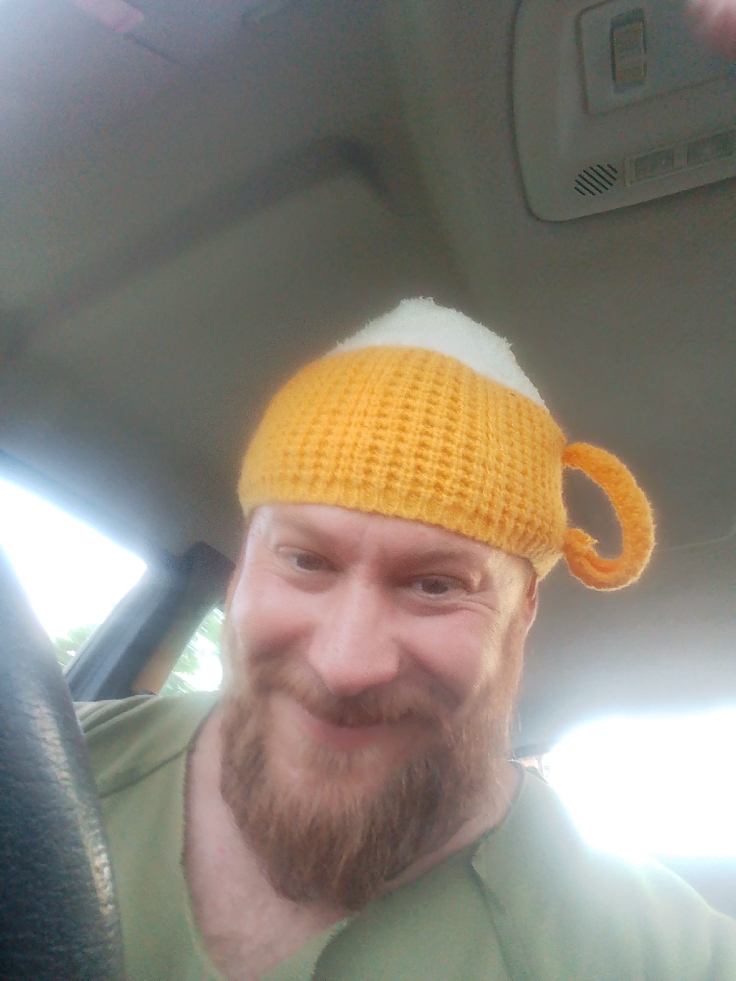 A close-up selfie of a man with a beard wearing a yellow and white knit hat resembling a beer mug. He is positioned behind the steering wheel inside a vehicle. The interior of the vehicle, including the ceiling and dashboard, is visible. The location is inside a car, the exact city is not discernible from the visual information provided.